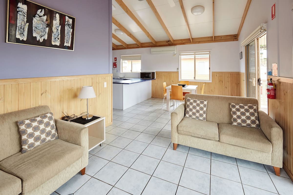 A cozy room with a sofa, a small table, and a lamp. There is a kitchen area in the background with a counter and chairs.