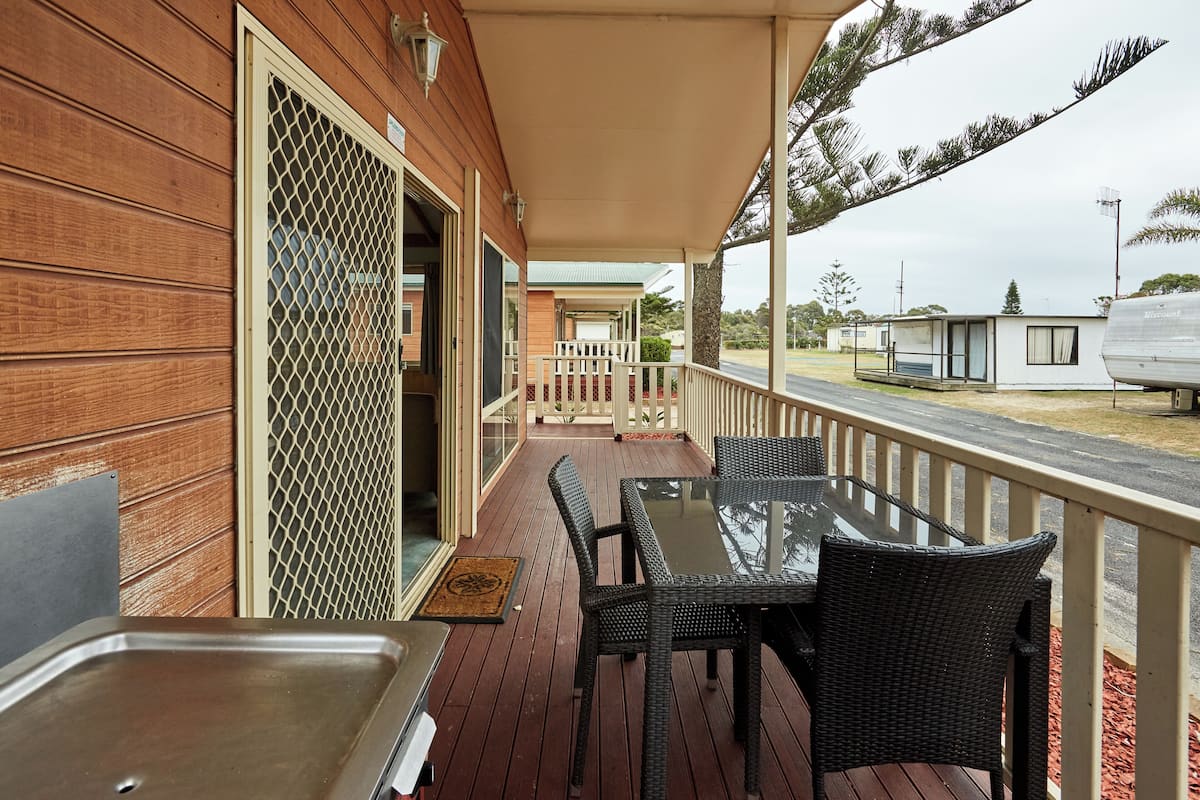 A wooden deck with a glass table and chairs, a metal sink, and a covered area with a railing.