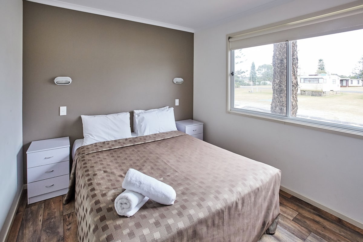 A neatly made bed with a patterned bedspread and two pillows, a bedside table with a lamp, and a window with a view of a tree and a trailer.