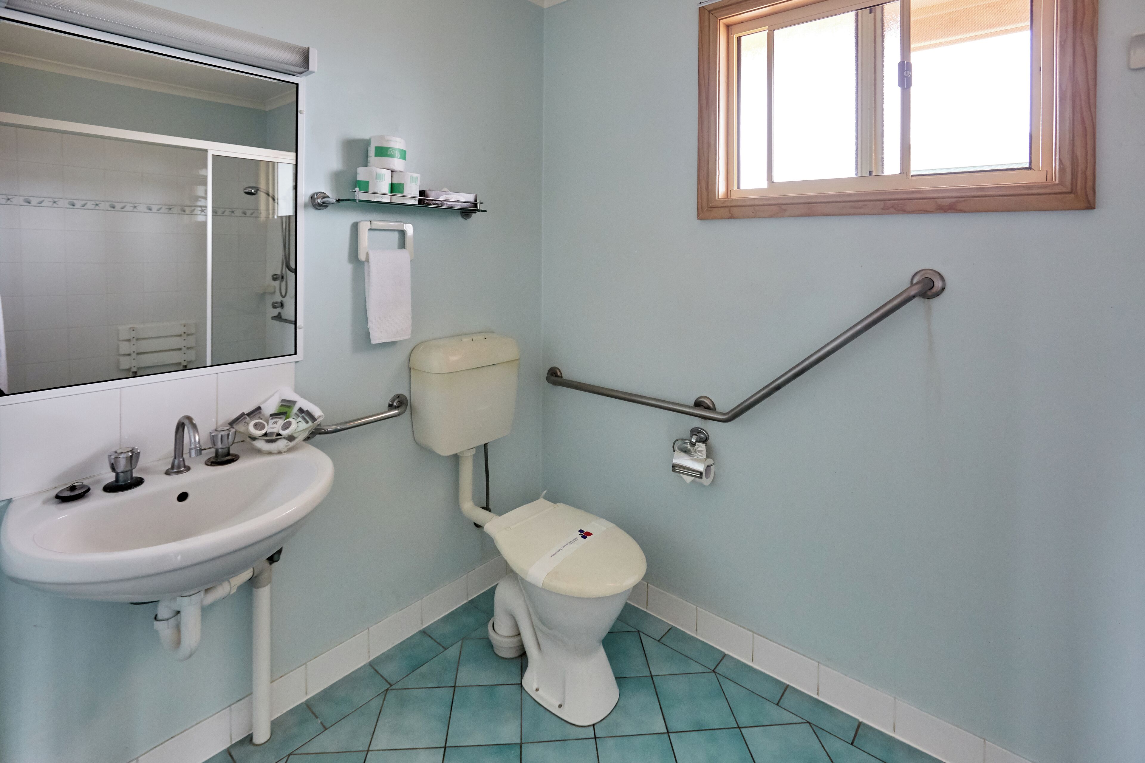 A bathroom with a white sink, toilet, and shower. There is a window above the toilet and a shelf with toiletries.