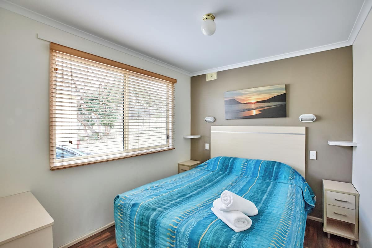 A bedroom with a large bed, a window with blinds, a bedside table, a nightstand, and a painting on the wall.