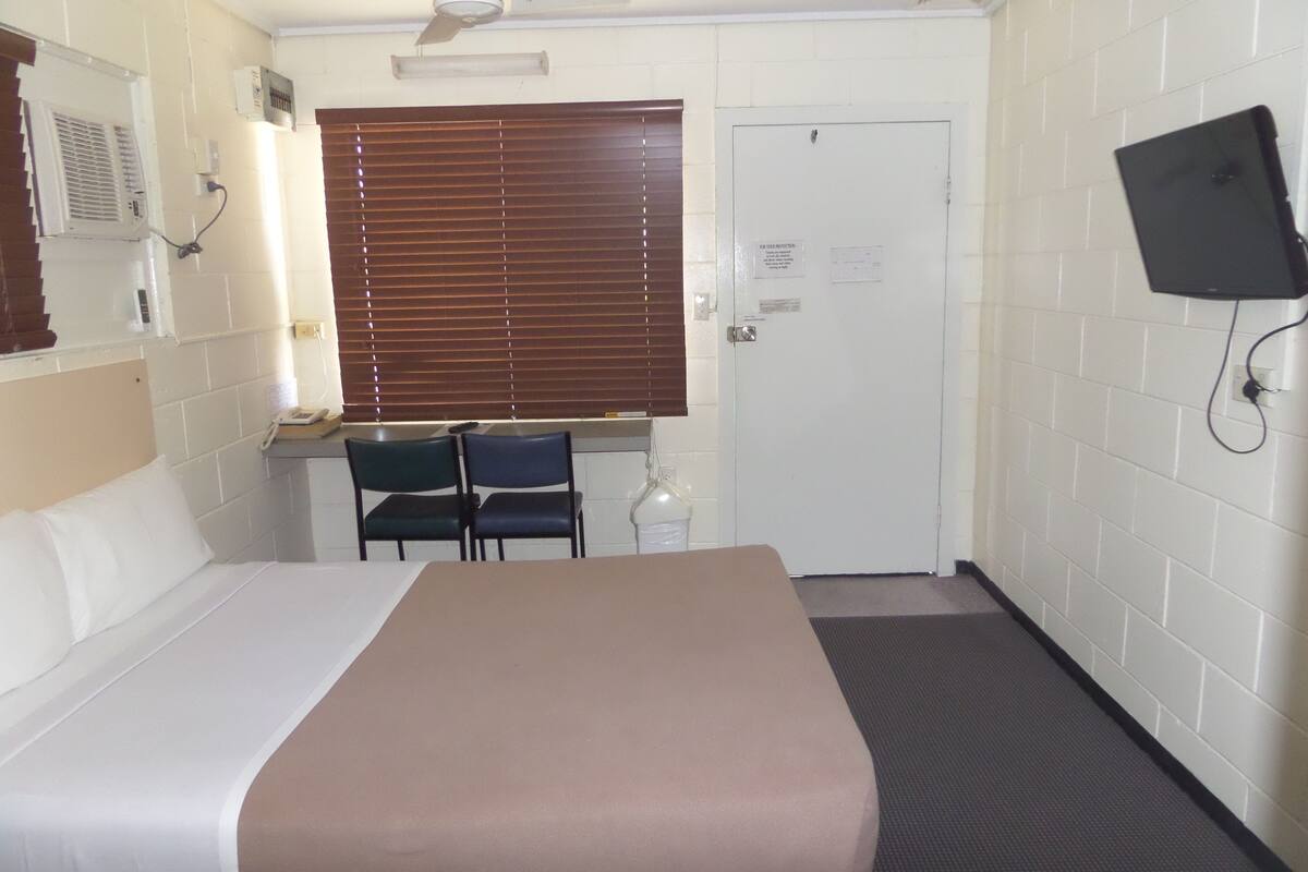 A hotel room with a bed, a desk with two chairs, a television mounted on the wall, and a door with a sign.