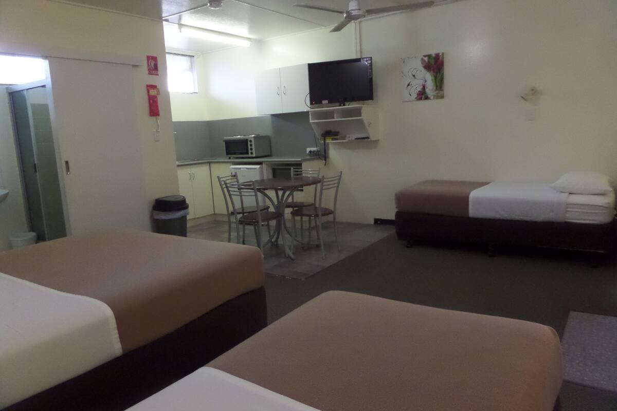 A hotel room with two beds, a small kitchenette, a dining table with chairs, and a TV mounted on the wall.