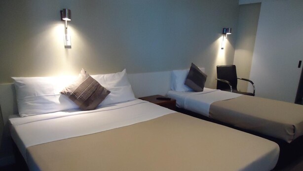 Quarto de hotel com duas camas de solteiro, cada uma com um travesseiro e um cobertor. Há duas mesas de cabeceira, uma com abajur e a outra com controle remoto.