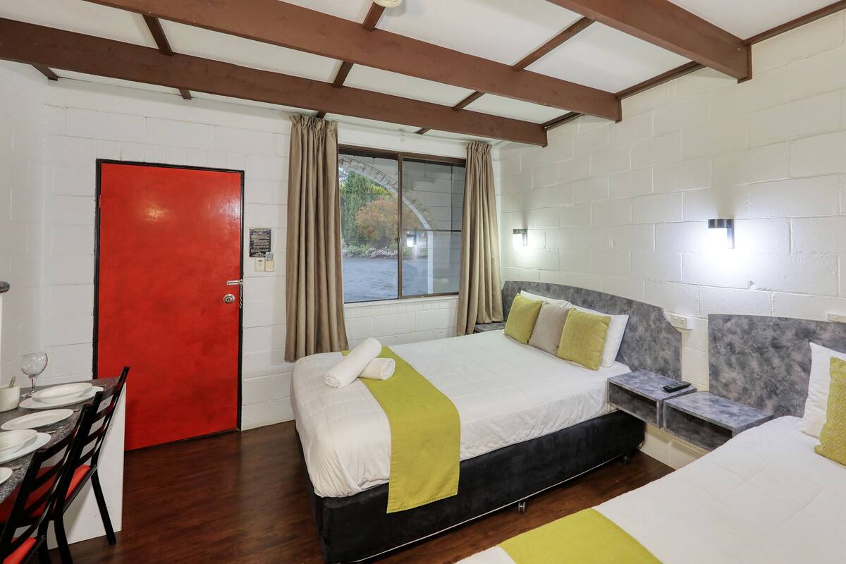 Een hotelkamer met een rode deur, een bed met een wit en geel sprei, een nachtkastje met een afstandsbediening, en een raam met gordijnen.