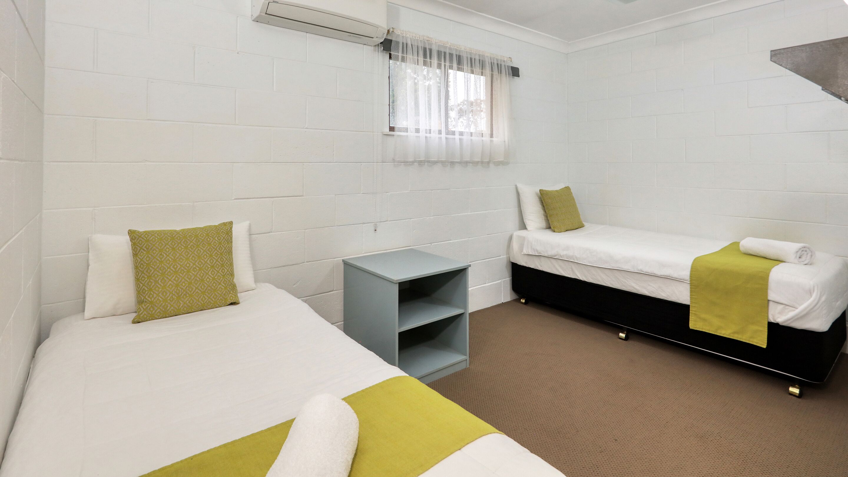 A hotel room with two beds, a small table, and a window with sheer curtains.