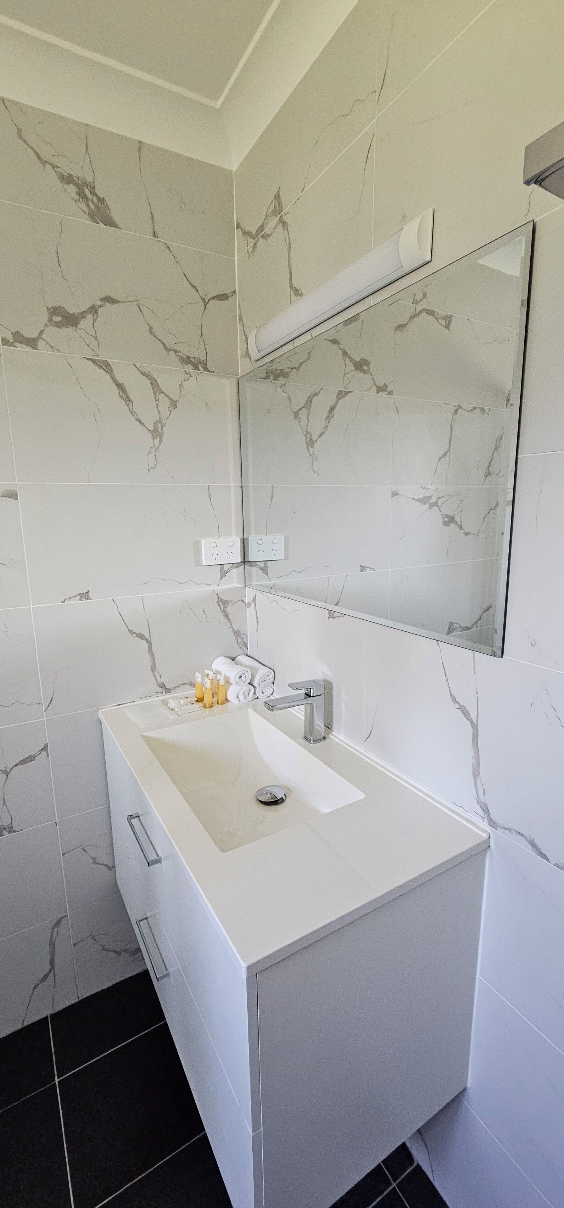Une salle de bain avec un mur aux motifs de marbre, un lavabo blanc avec un miroir au-dessus, et un sol carrelé de noir.