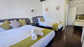A hotel room with two beds, white linens, green and yellow pillows, a small bedside table, and a white brick wall.