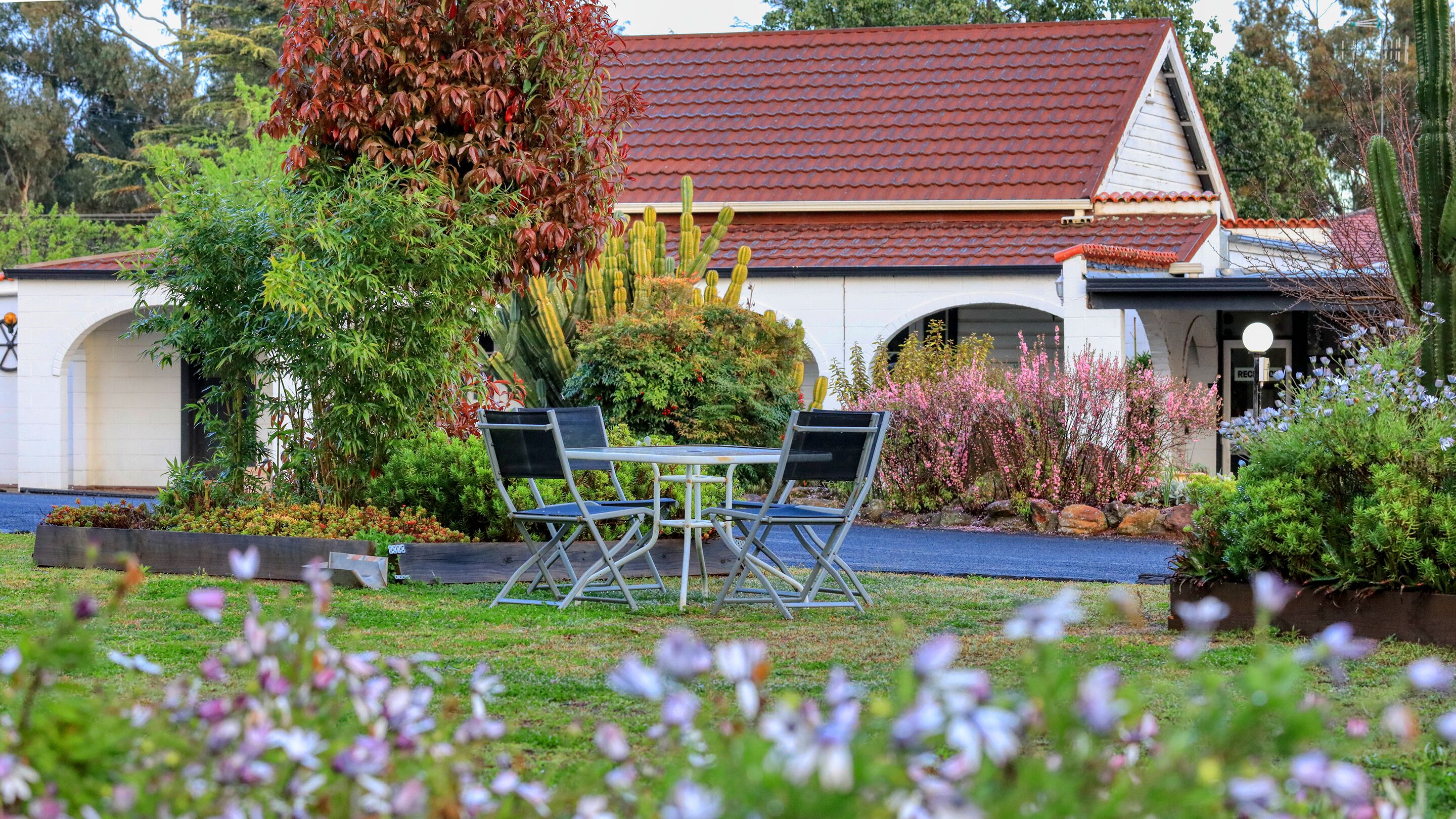 Un jardín con una mesa y sillas, un edificio de techo rojo y diversas plantas y flores.