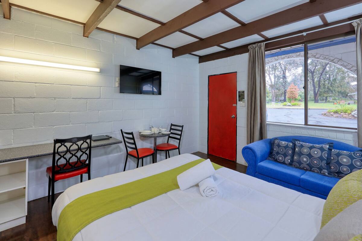Een hotelkamer met een bed, een bureau met stoelen, een rode deur en een groot raam met uitzicht op bomen en een parkeerplaats.