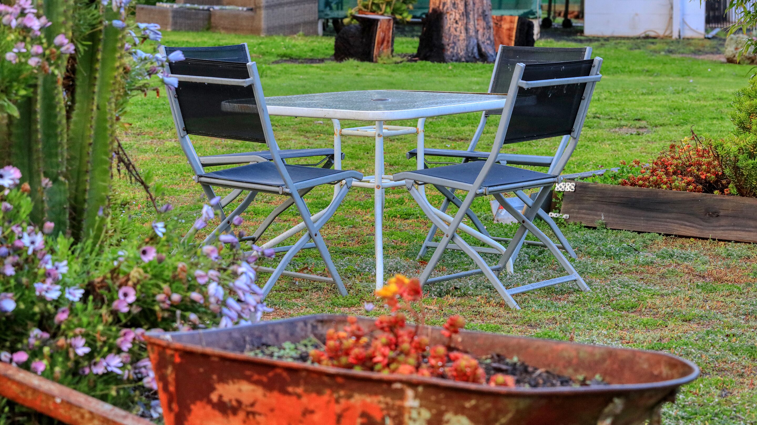 A metal table and chairs in a garden with a wheelbarrow in the foreground.