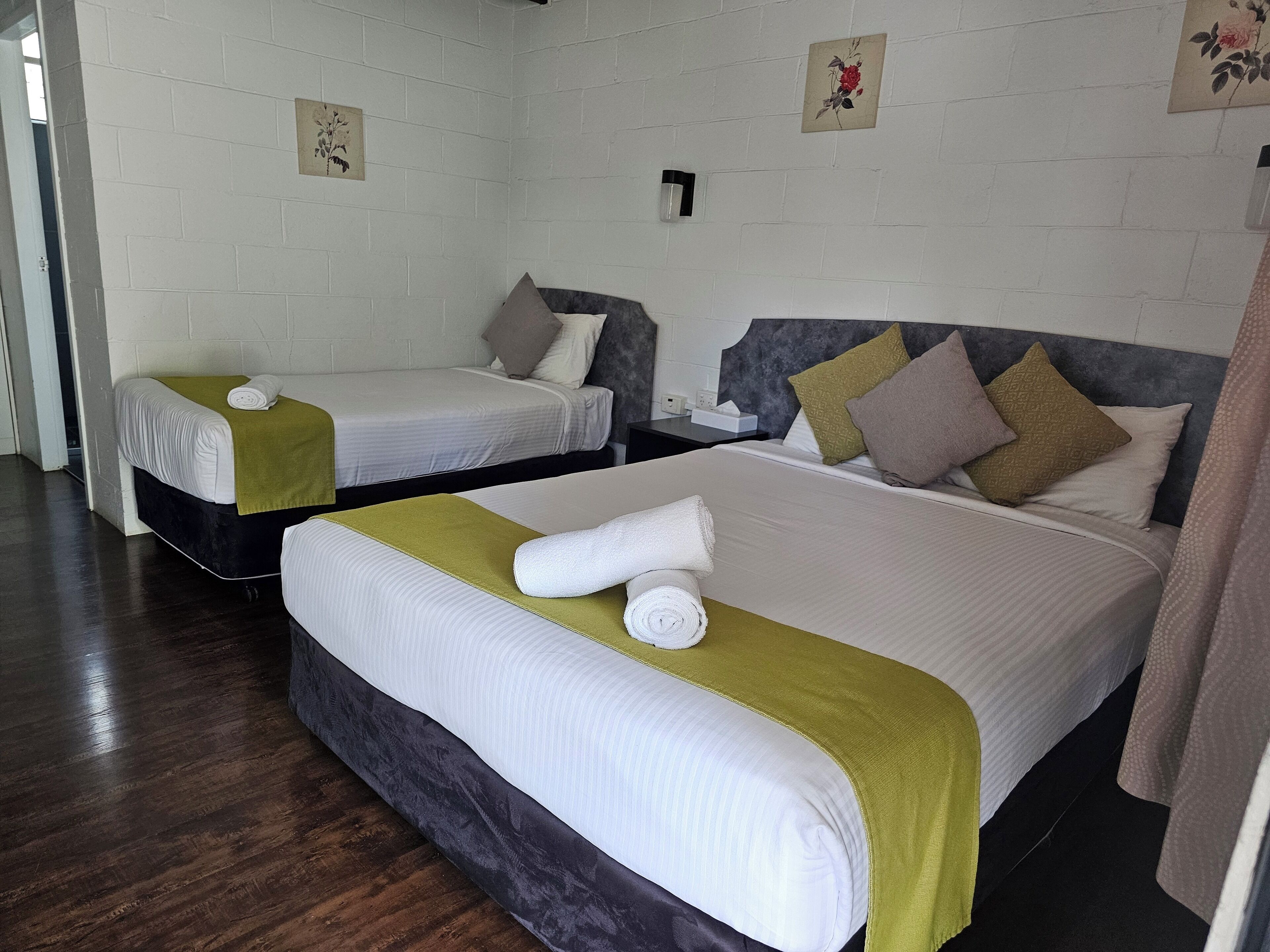 A hotel room with two beds, each with white linens and green throw pillows. There is a small bedside table with a lamp and a tissue box. The room has white brick walls and a wooden floor.