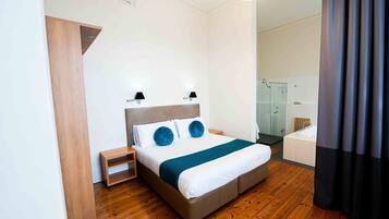 A modern hotel room with a wooden floor, a large bed with a headboard, bedside tables, a nightstand, a built-in shower, and a bathroom with a bathtub.