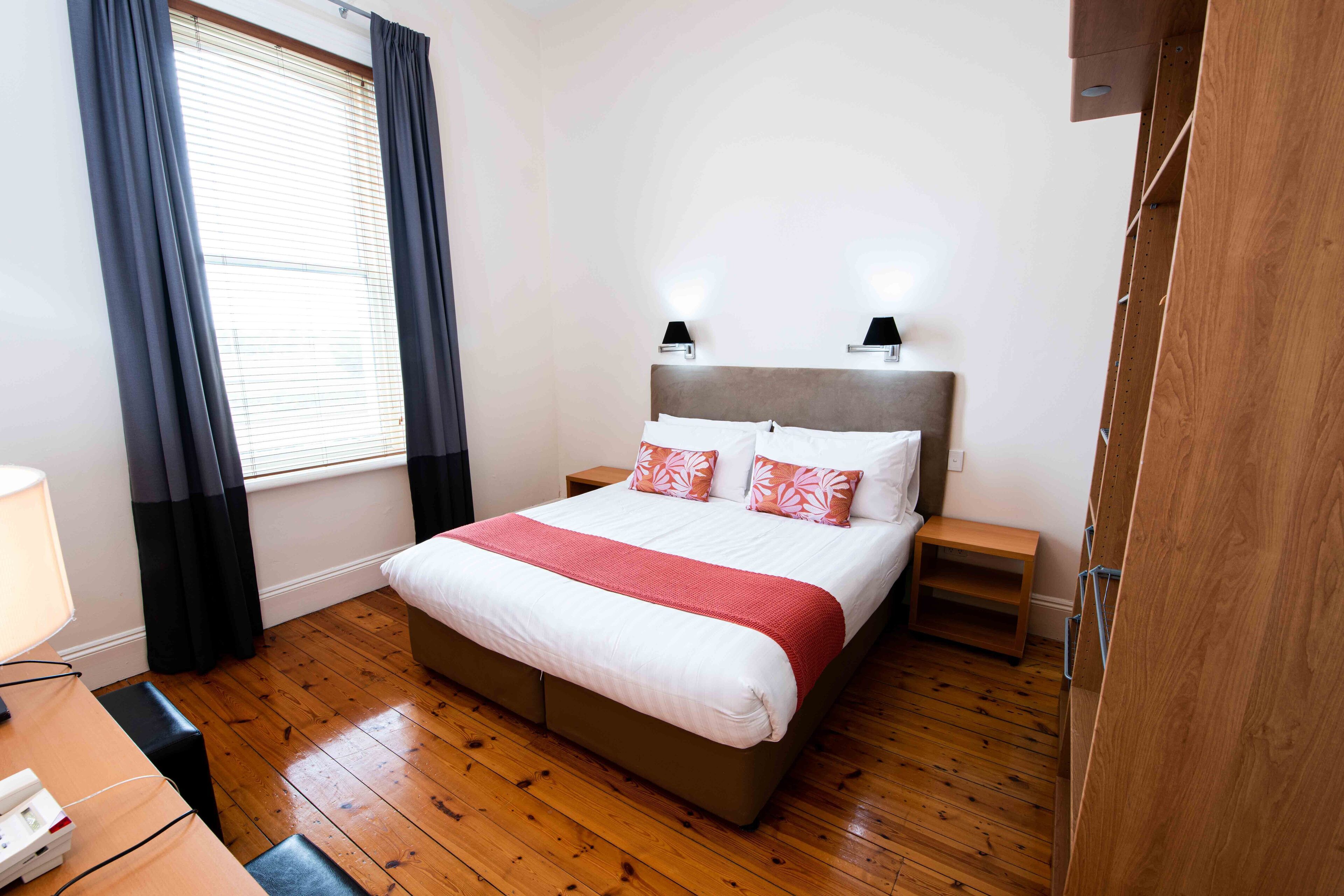 A hotel room with a wooden floor, a bed with white and red bedding, two bedside tables with lamps, and a window with curtains.