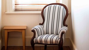 Une pièce d’angle avec un plancher en bois, une chaise à rayures, une petite table en bois et une fenêtre avec des stores.