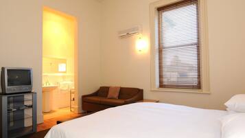 Habitación de hotel con cama, televisor en un mueble bajo, sofá, ventana con persianas y equipo de aire acondicionado.