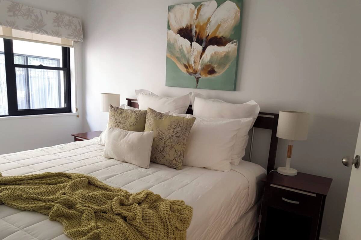 Uma cama bem arrumada com um cobertor verde de malha, um quadro floral acima e um abajur em uma mesa de cabeceira.