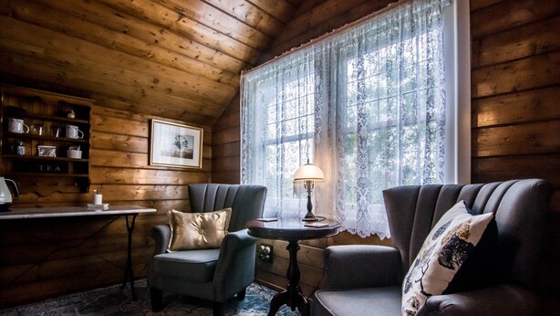 A cozy room with wooden walls, two upholstered chairs, a small table with a lamp, a framed picture on the wall, and a shelf with tea items. - Heytesbury House (Cobden)