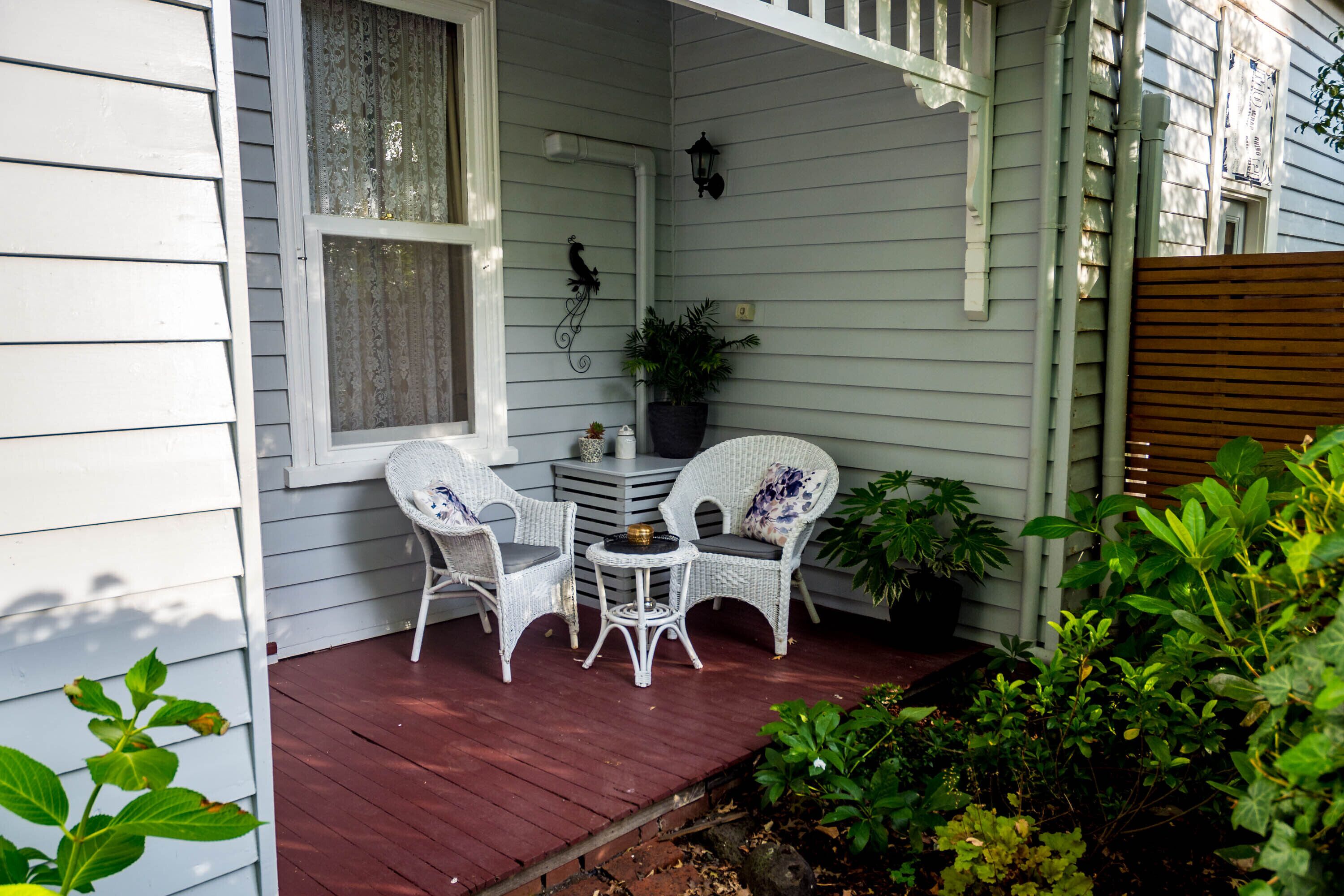 A porch with white wicker chairs and a small table, surrounded by plants and a wooden fence.