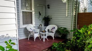 A porch with white wicker chairs and a small table, surrounded by plants and a wooden fence.