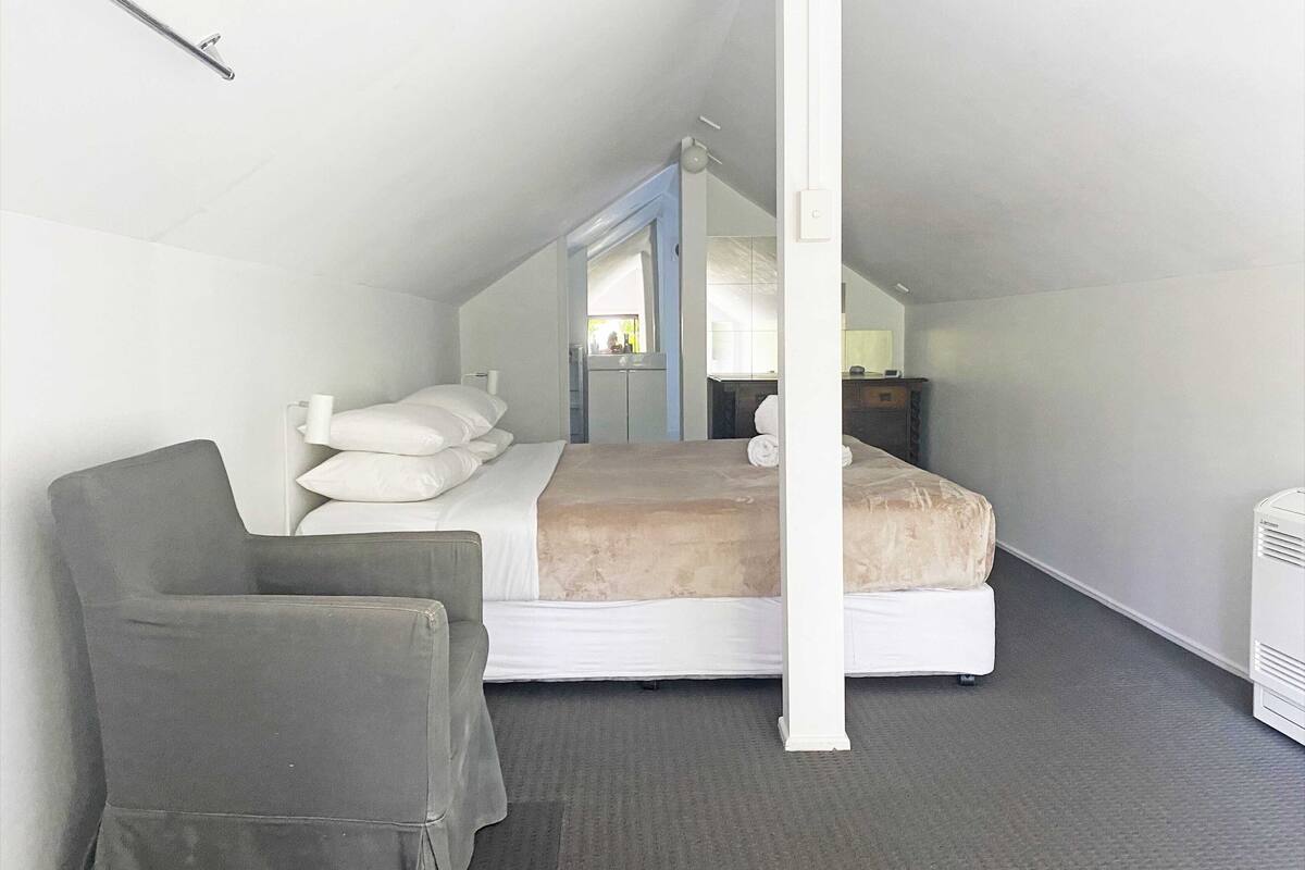 A small, single-room hotel with a sloped ceiling, a bed with white linens, a grey armchair, a small desk with a lamp, and a white air conditioning unit.