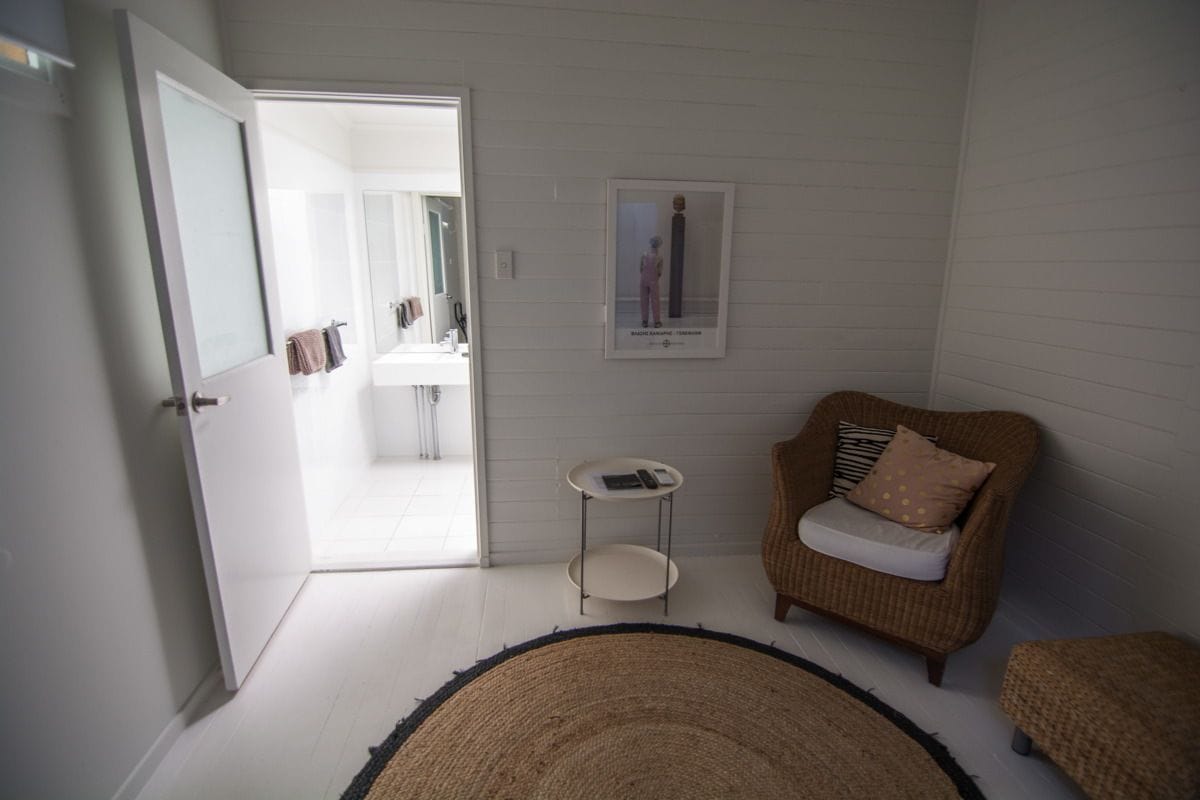 Une pièce comprenant un fauteuil en osier, une petite table ronde et une porte donnant sur une salle de bain.