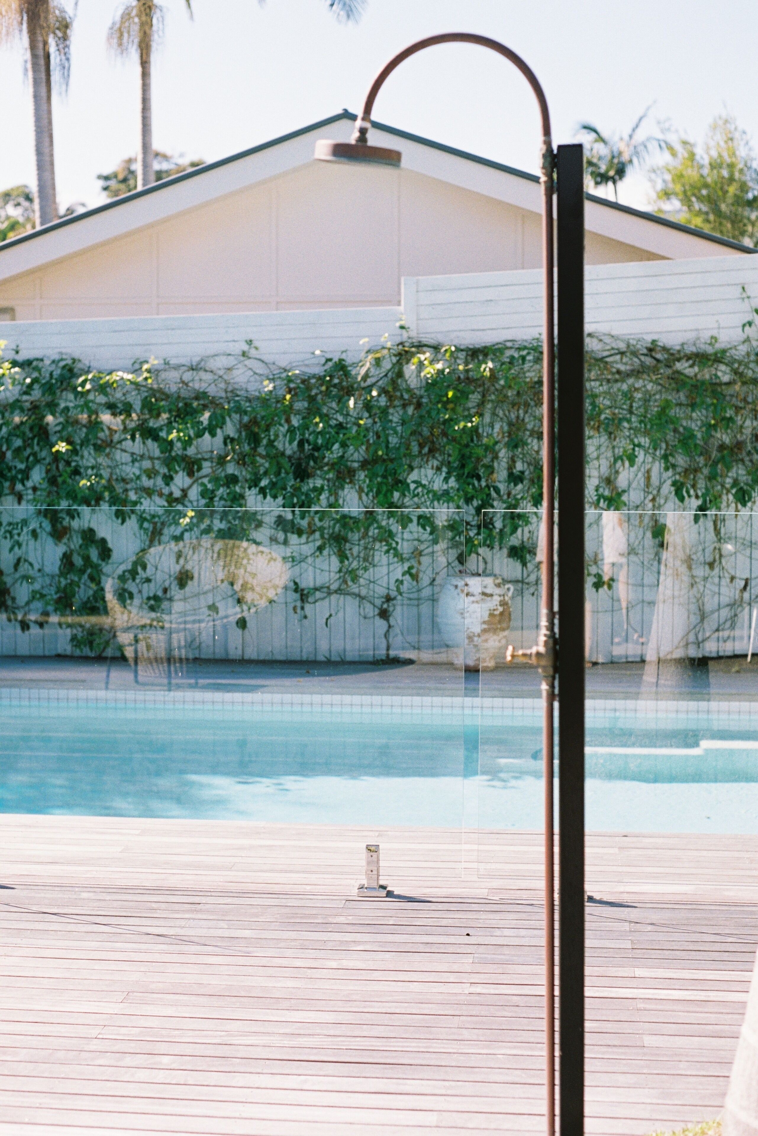 Une piscine dotée d’une terrasse en bois, d’une structure métallique et d’un bâtiment à l’extérieur blanc, avec un feuillage verdoyant en arrière-plan.