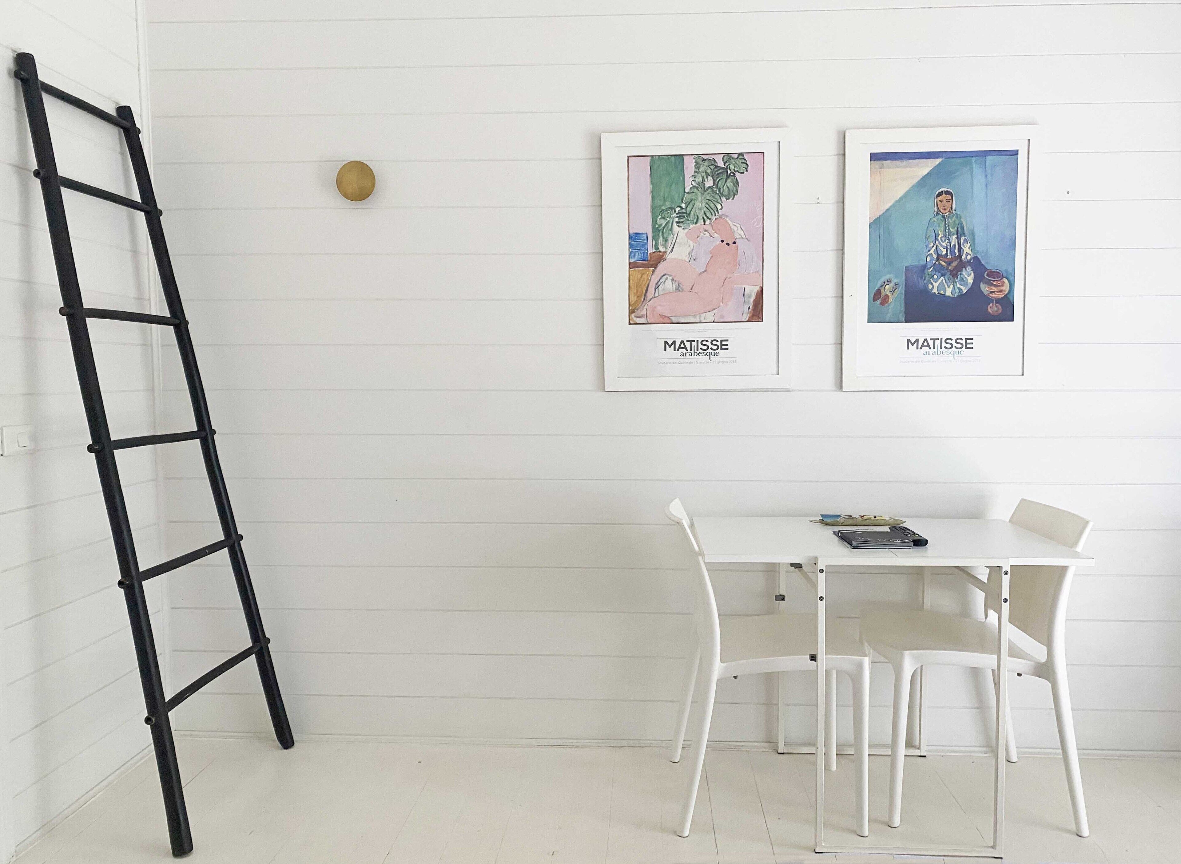 A white room with a white table and chairs, a black ladder, and two framed artworks on the wall.
