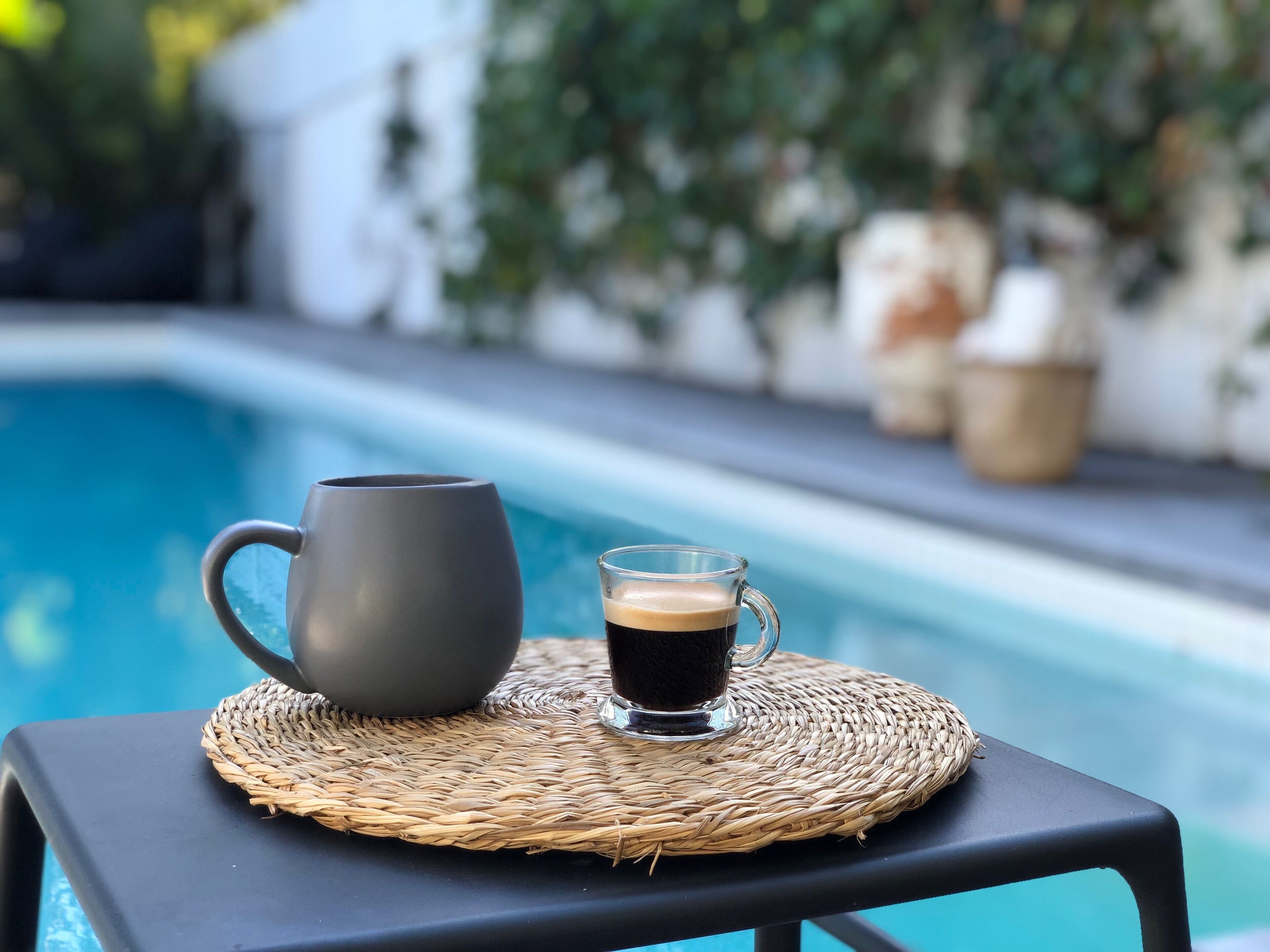 A poolside setting with a dark-colored mug and a glass of dark liquid, possibly coffee, on a woven mat.
