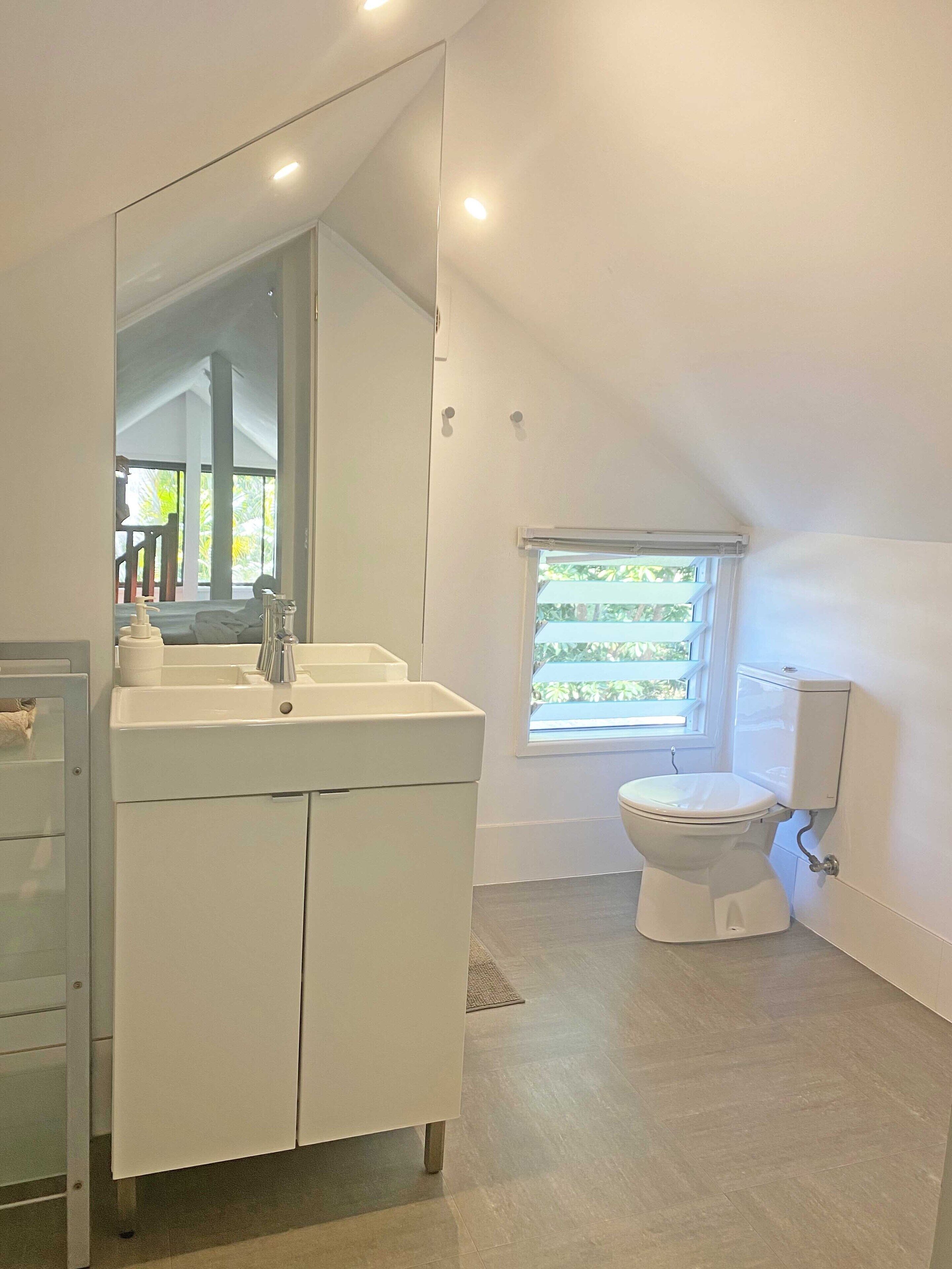 A modern bathroom with a white vanity, a large mirror, a toilet, and a window with a view of greenery.
