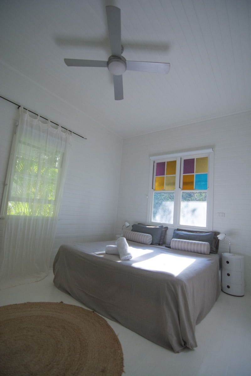 A bedroom with a bed, a ceiling fan, a window with colorful panes, and a bedside lamp.