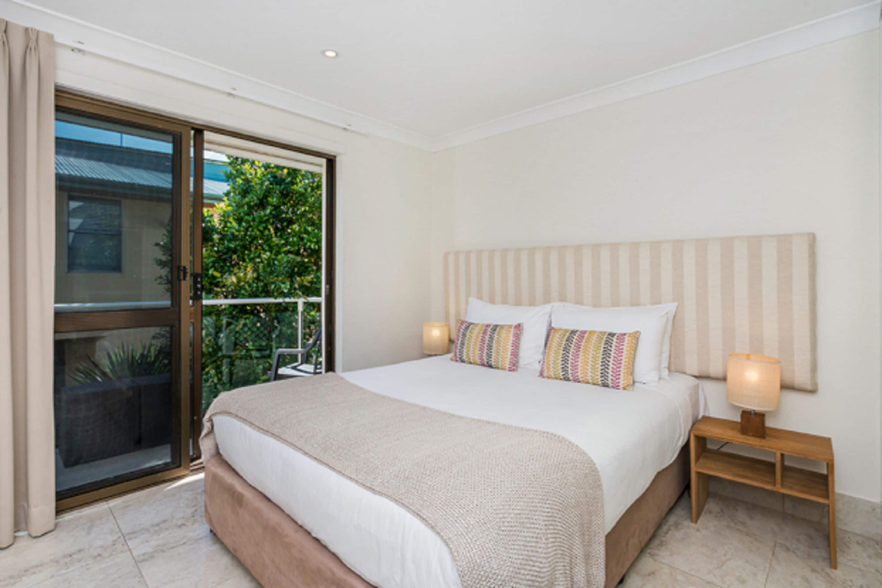 A bedroom with a large bed, a nightstand, a lamp, a sliding glass door to a balcony, and a view of greenery outside.