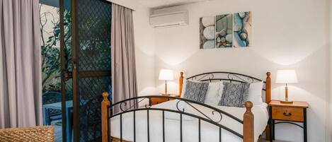 A bedroom with a bed, two bedside tables, a window with curtains, and a wall-mounted air conditioner.