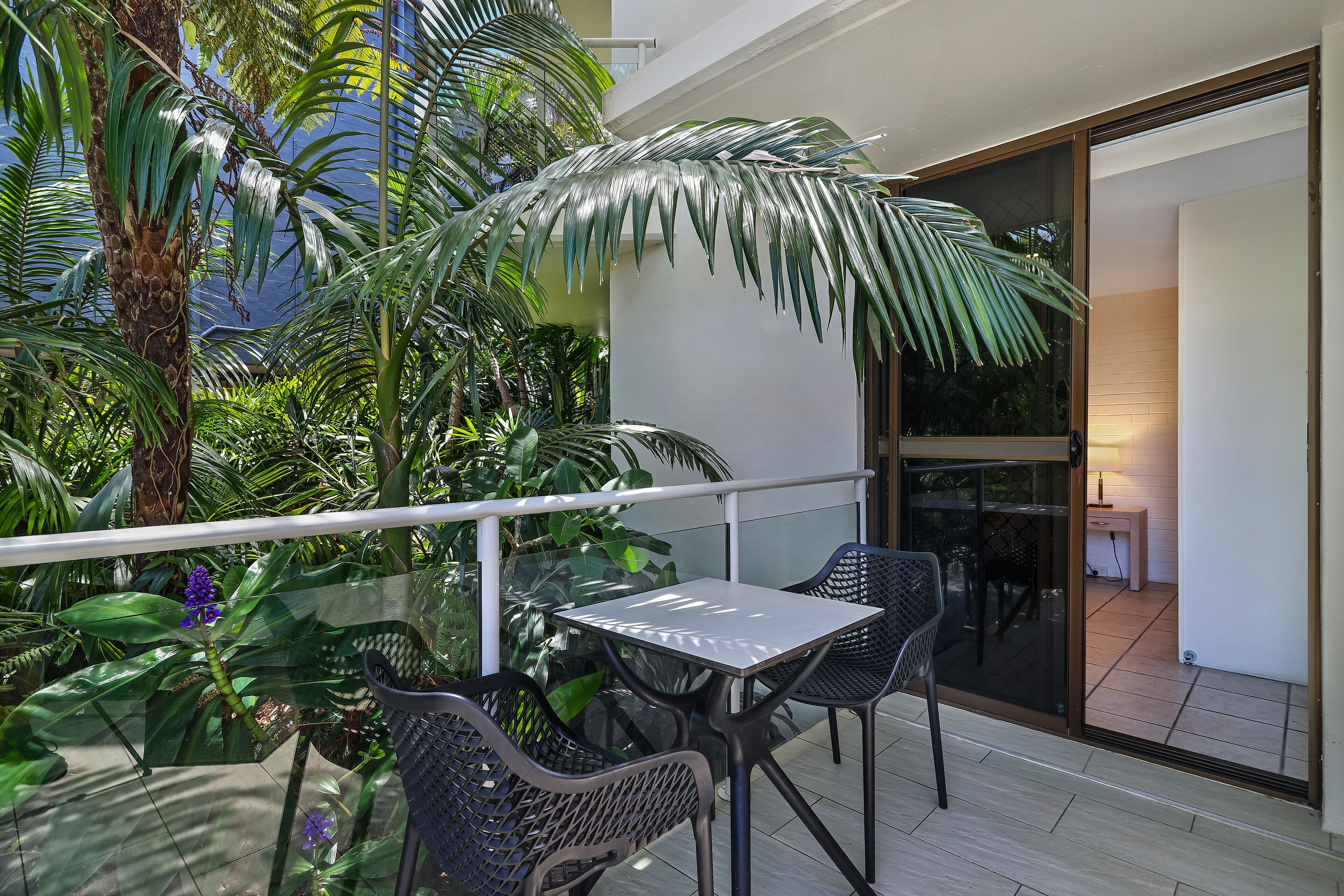 A balcony with a glass table and chairs, surrounded by lush greenery and a view of the interior.