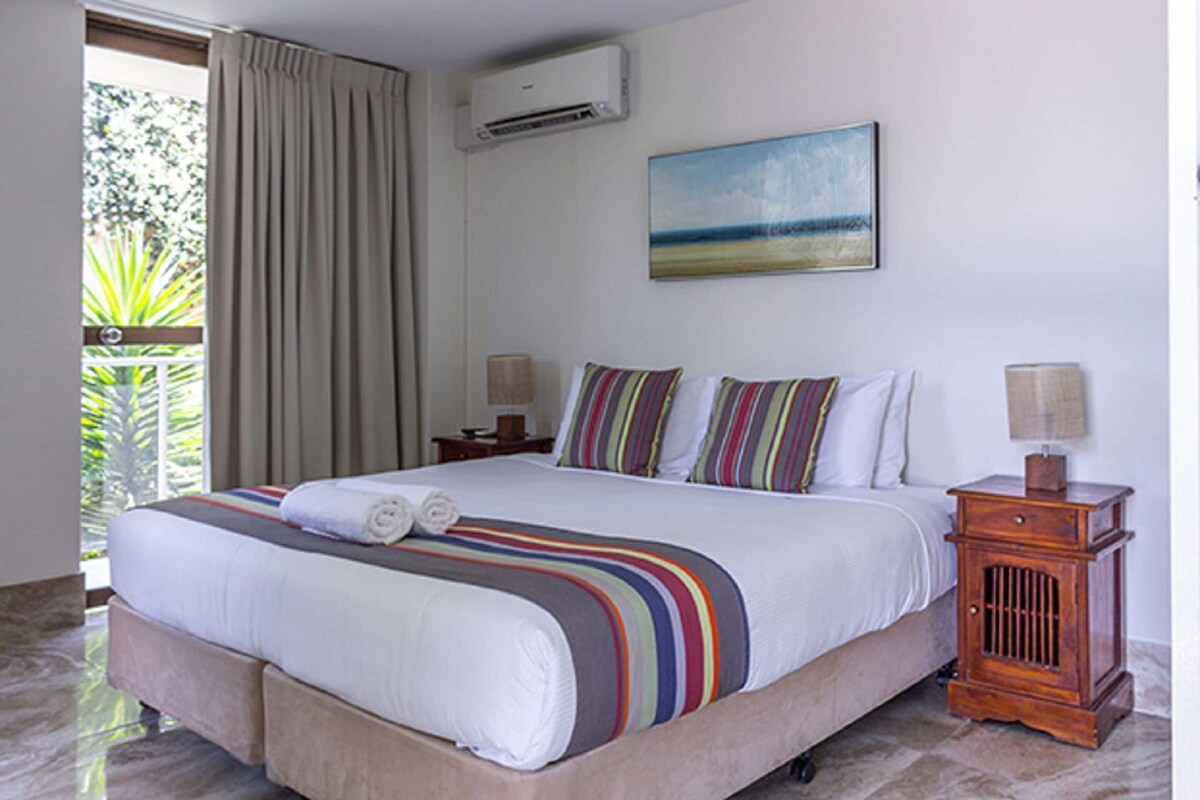 A hotel room with a bed, a nightstand, a lamp, an air conditioner, and a painting on the wall.