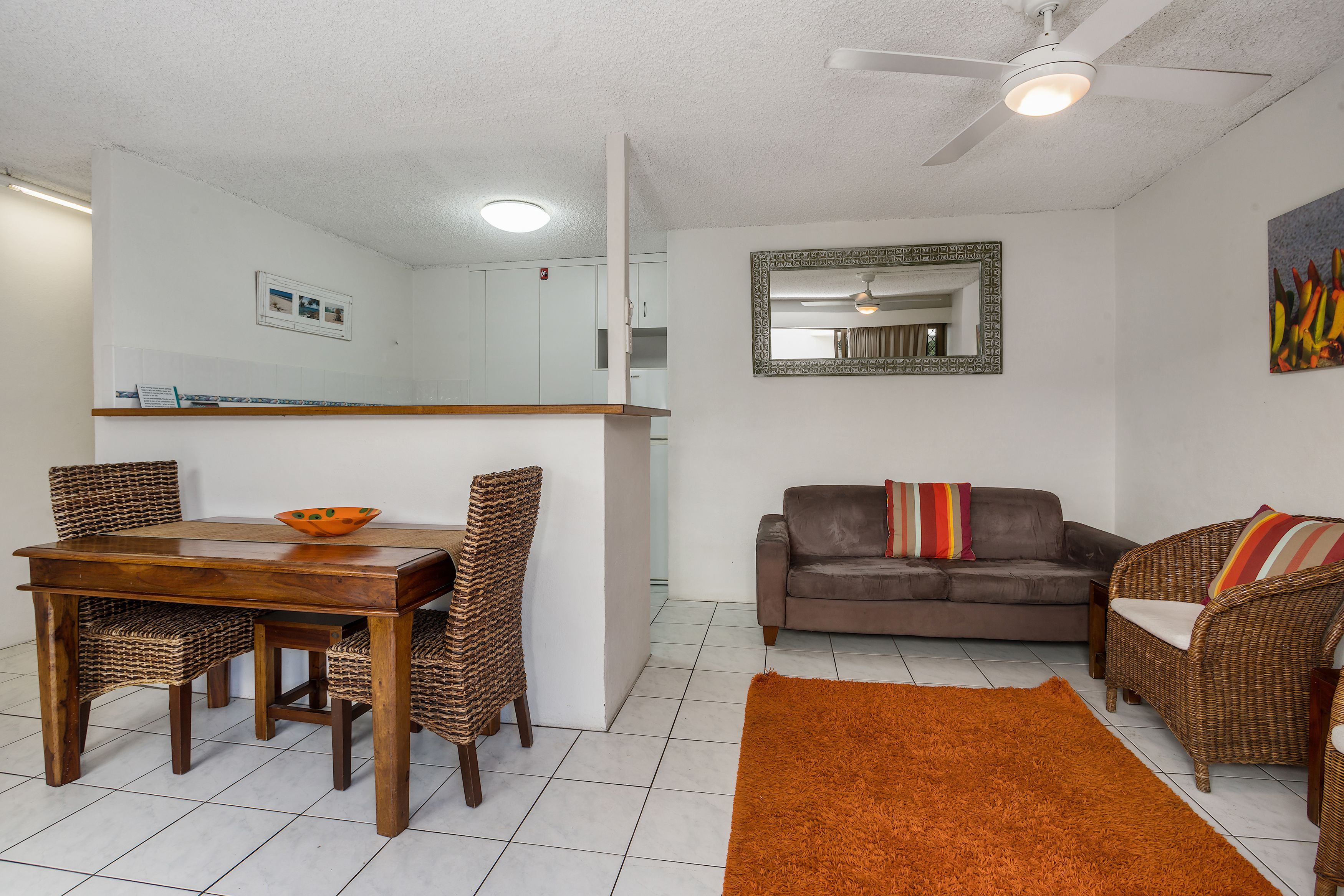 A dining area with a wooden table and wicker chairs, a brown sofa with colorful cushions, and a brown rug.