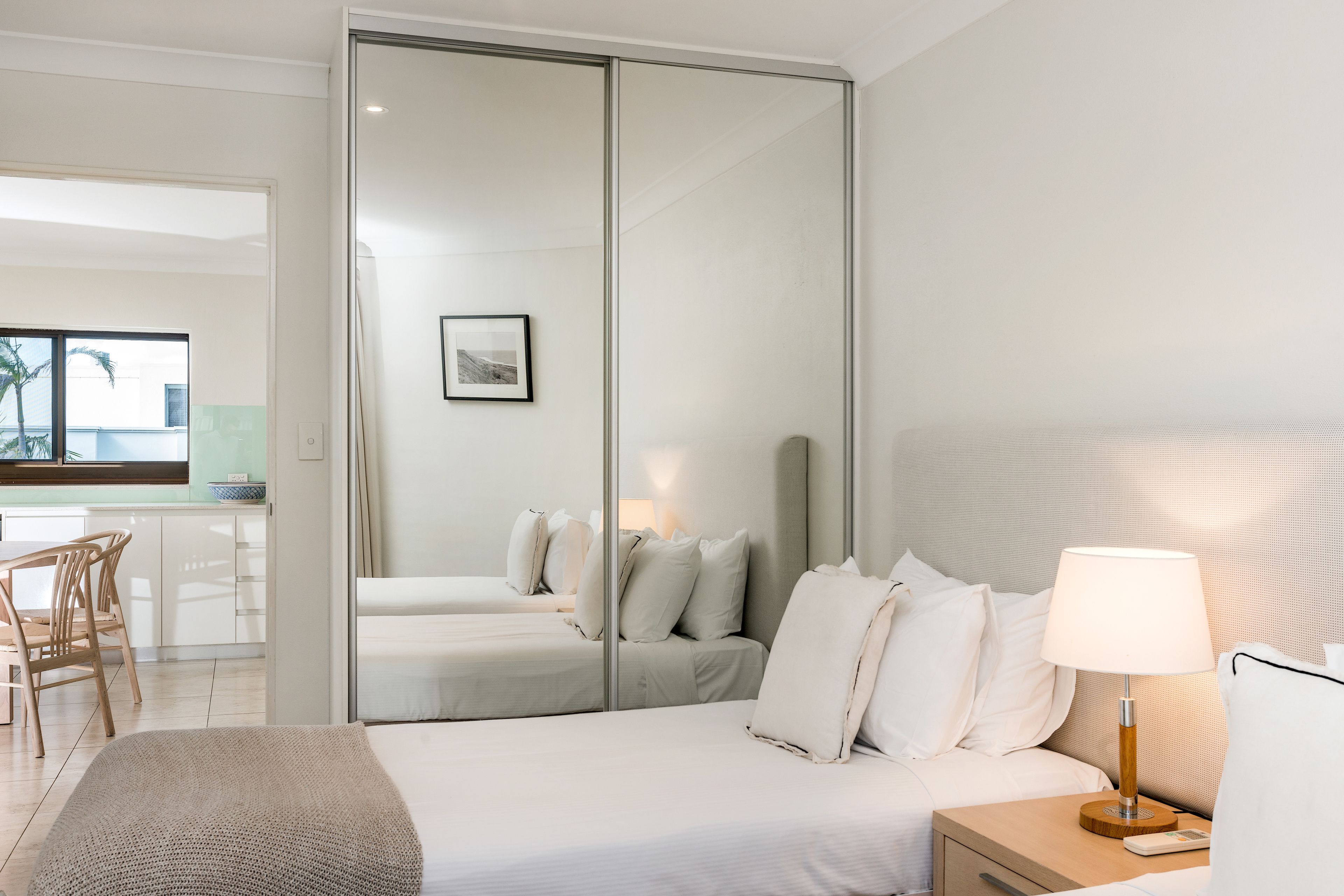 A modern hotel room with a large bed, a bedside table with a lamp, a chair, and a view of the kitchen and balcony.