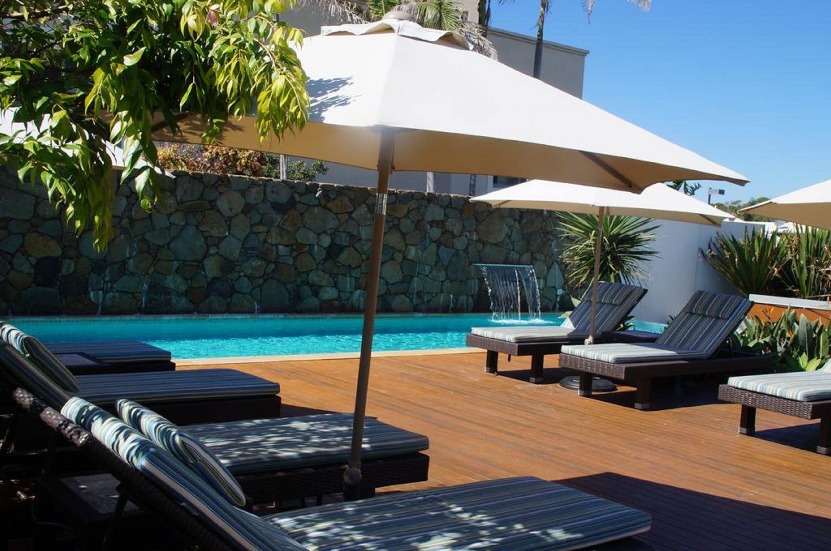 A poolside area with lounge chairs and umbrellas, a stone wall, and a clear blue pool.