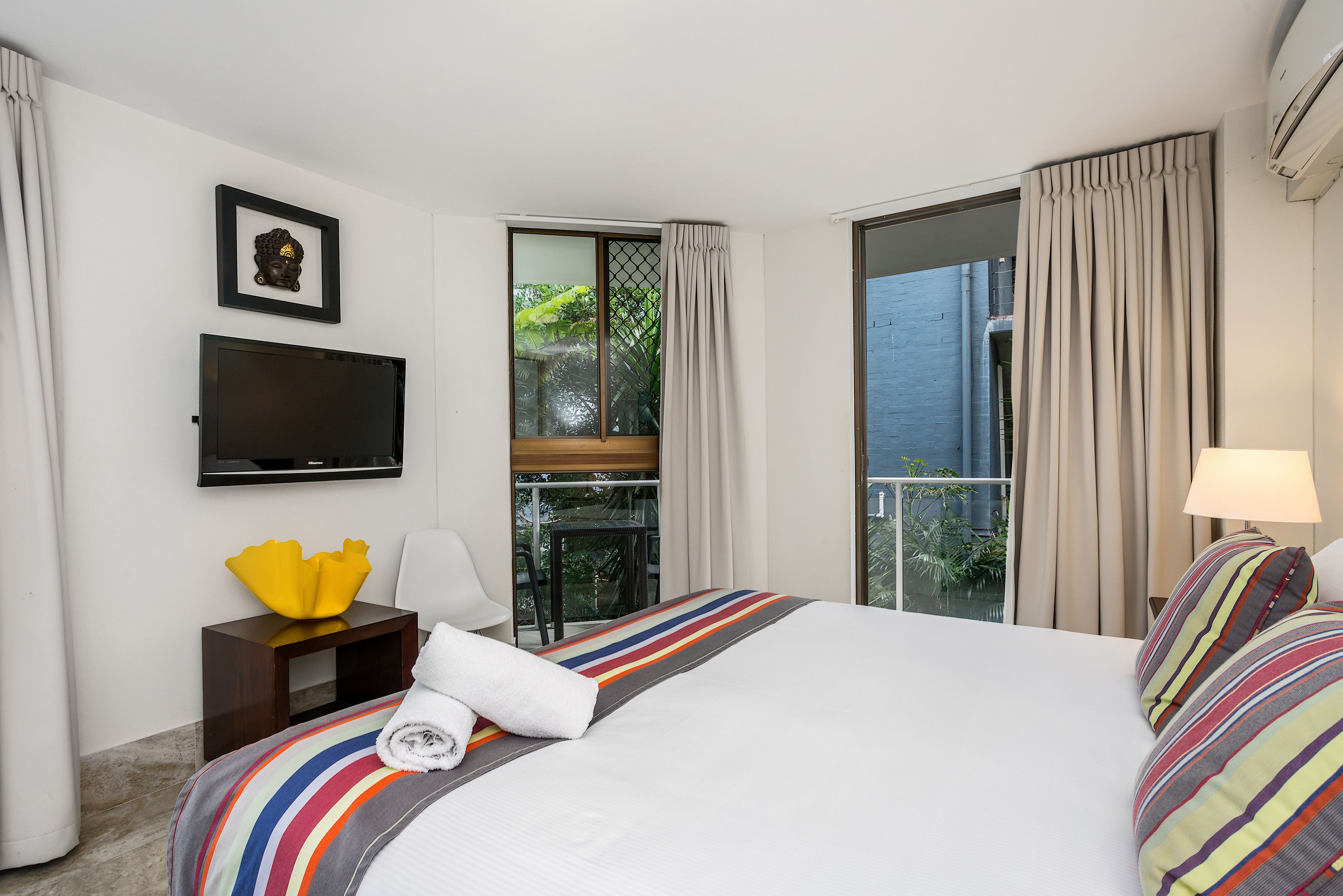 A hotel room with a bed, a TV, a small table with a yellow vase, a balcony with curtains, and a view of greenery.