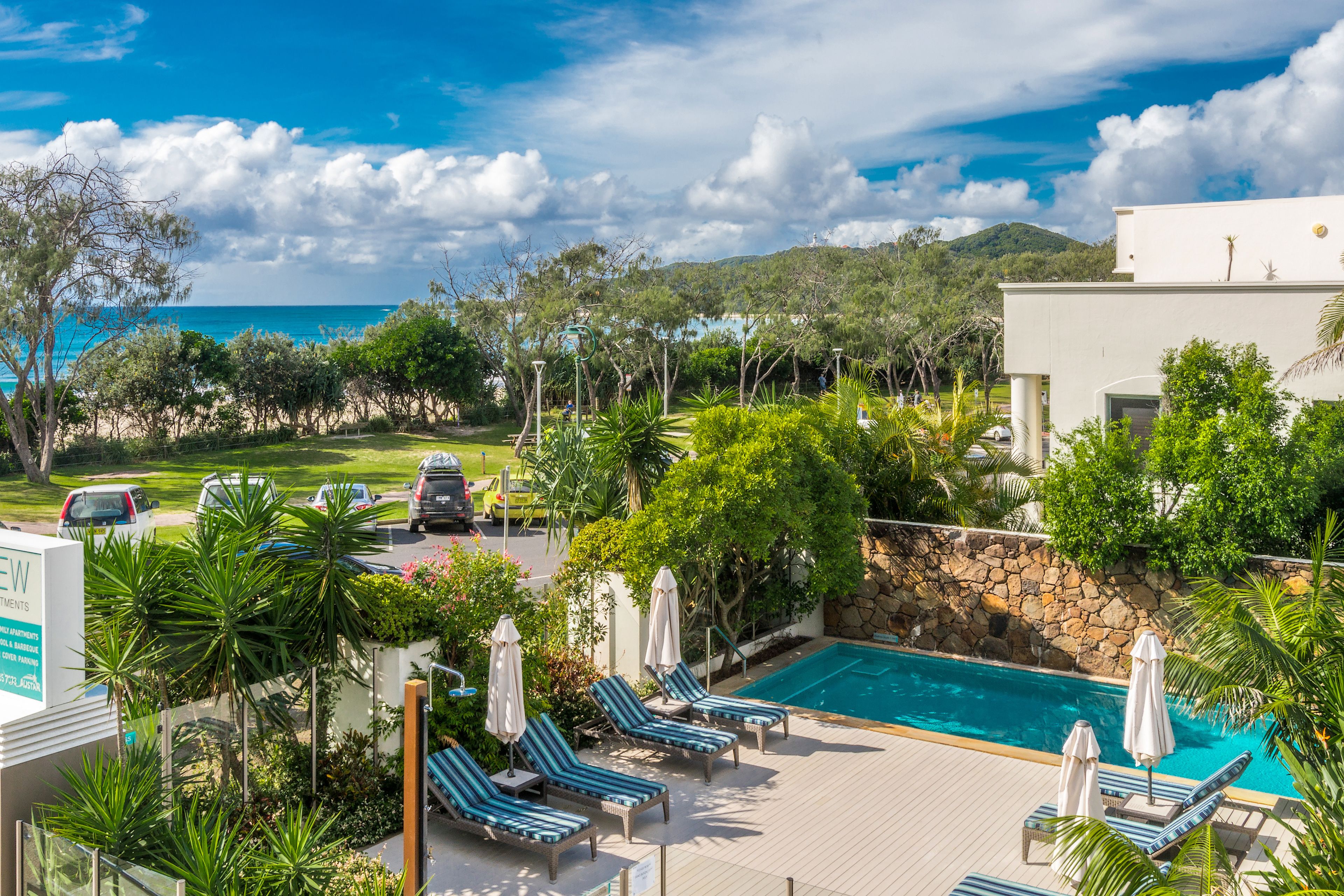 A pool area with lounge chairs and umbrellas, surrounded by greenery and a view of the ocean.