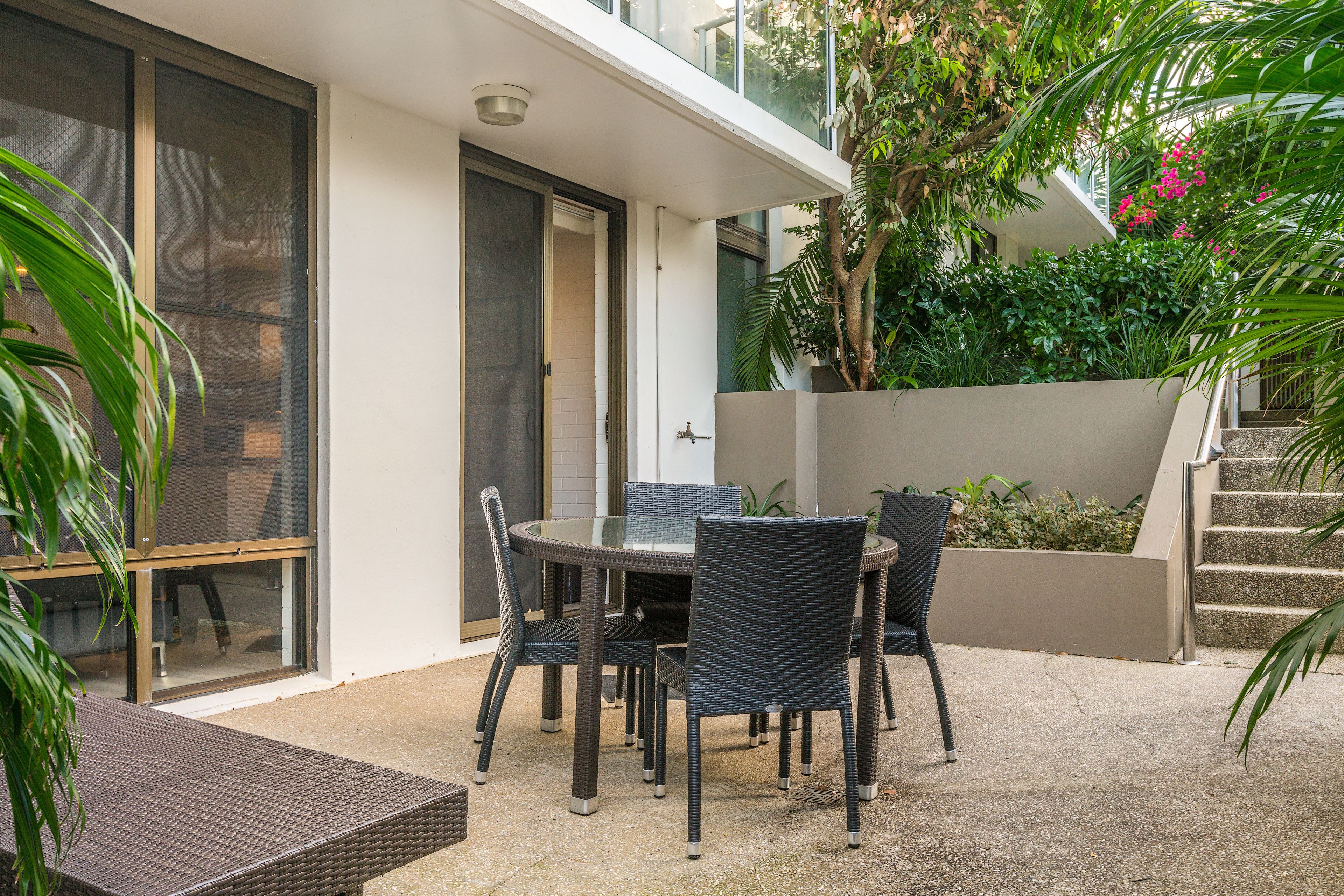 A patio area with a round table and chairs, a glass door leading to a covered entrance, and a staircase with a concrete wall and greenery.