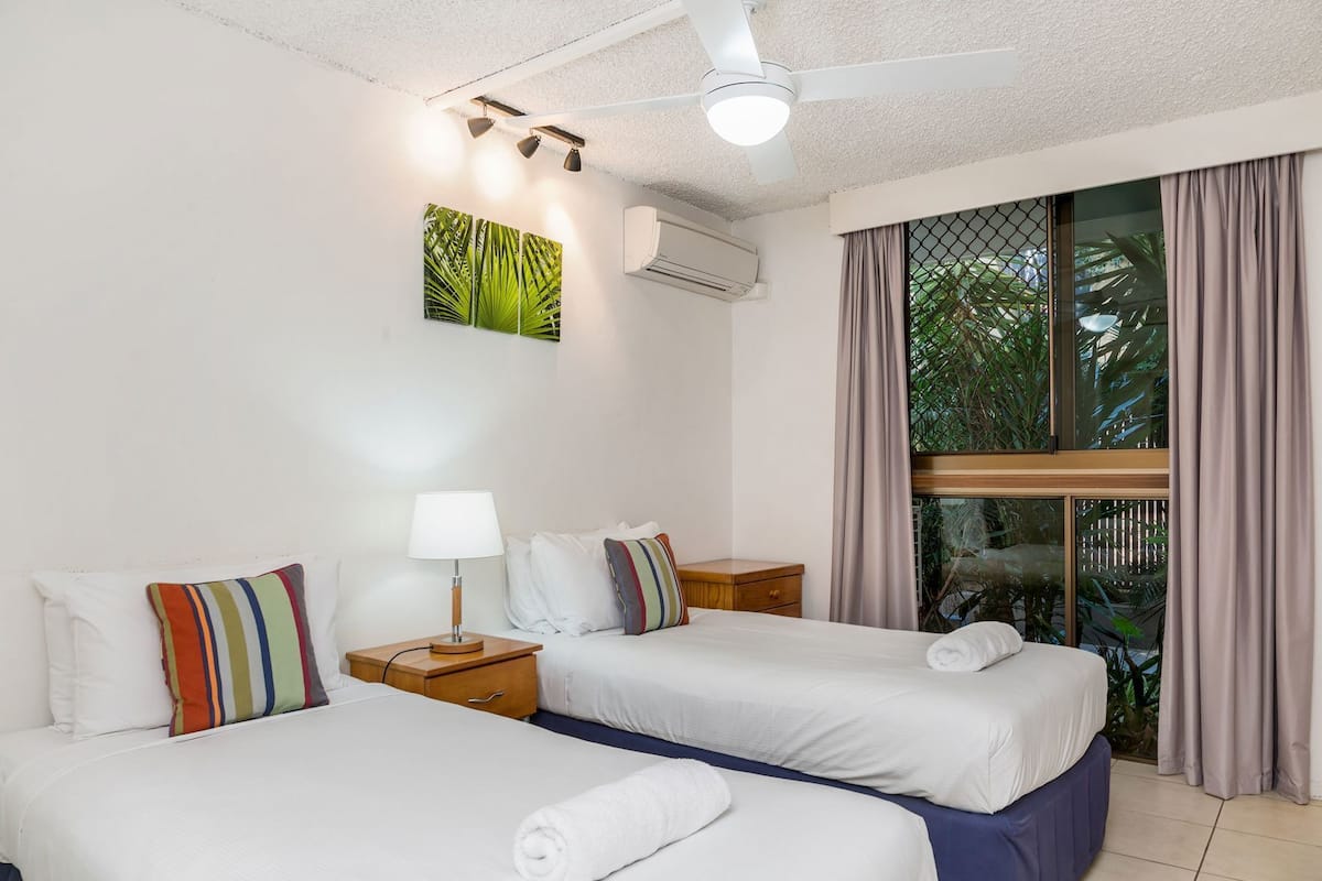 Ein Hotelzimmer mit zwei Betten, einem Deckenventilator, einer Klimaanlage, einem Fenster mit Vorhängen und einer durch das Fenster sichtbaren Pflanze.