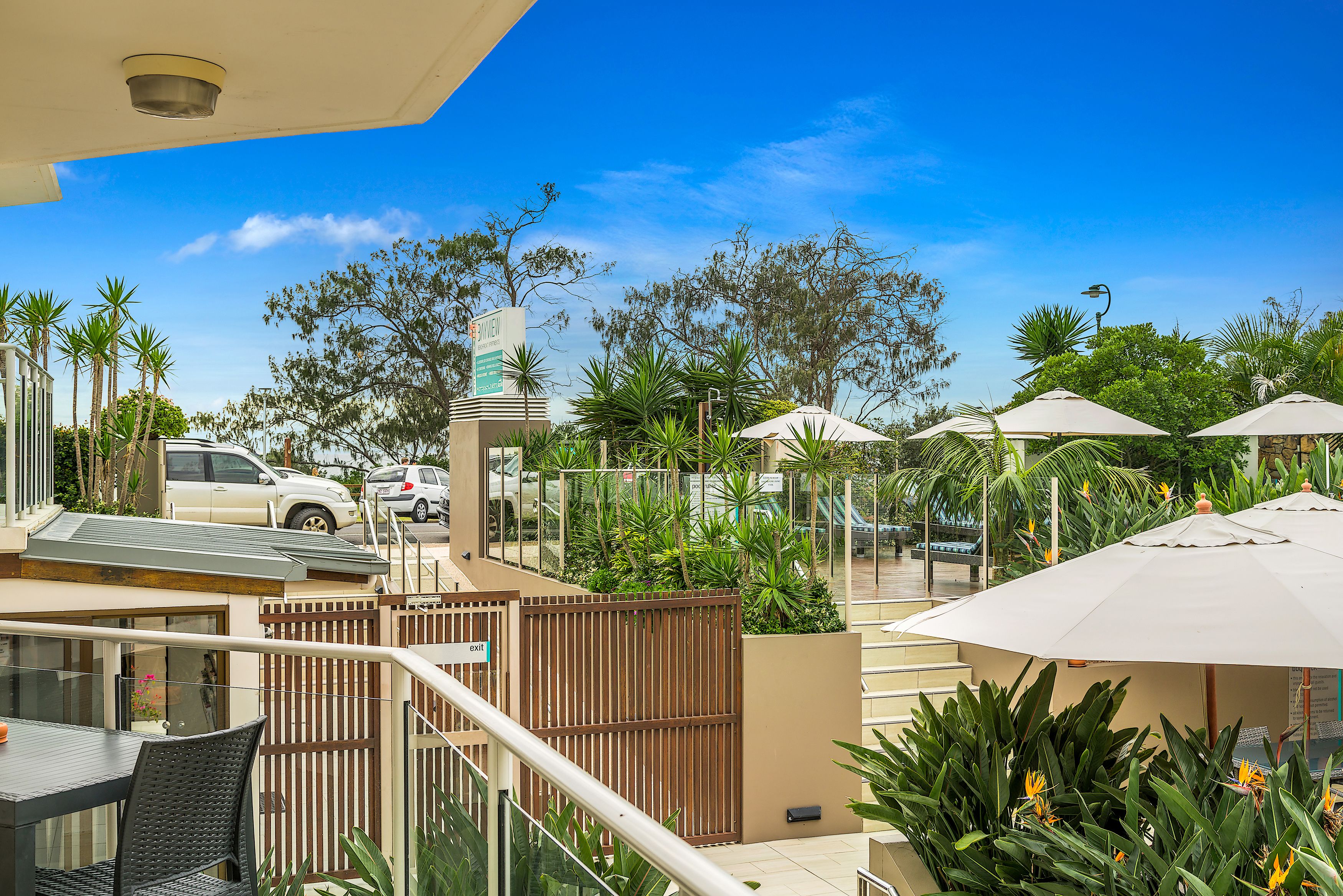 A hotel balcony with a view of parked cars, a gate, and outdoor umbrellas.