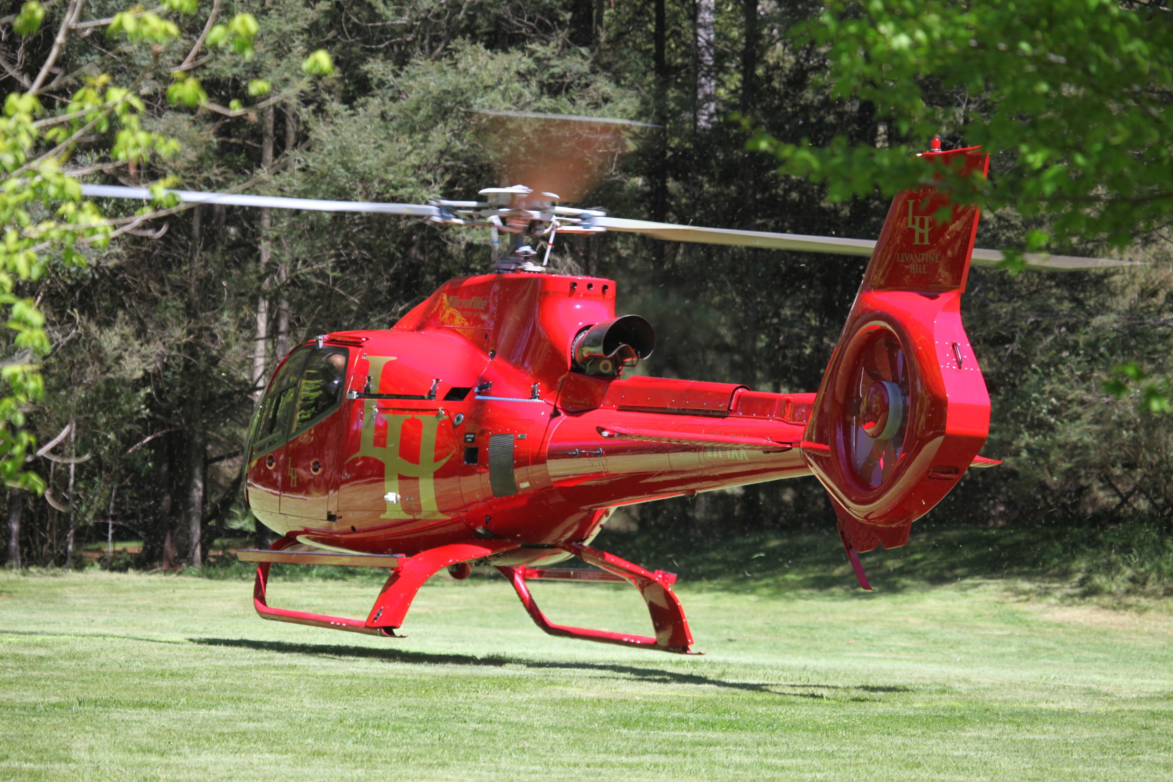 A red helicopter with a visible logo on its tail, parked on a grassy field with trees in the background.