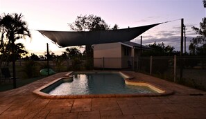 A swimming pool with a shaded area, surrounded by a brick patio, and a building in the background.