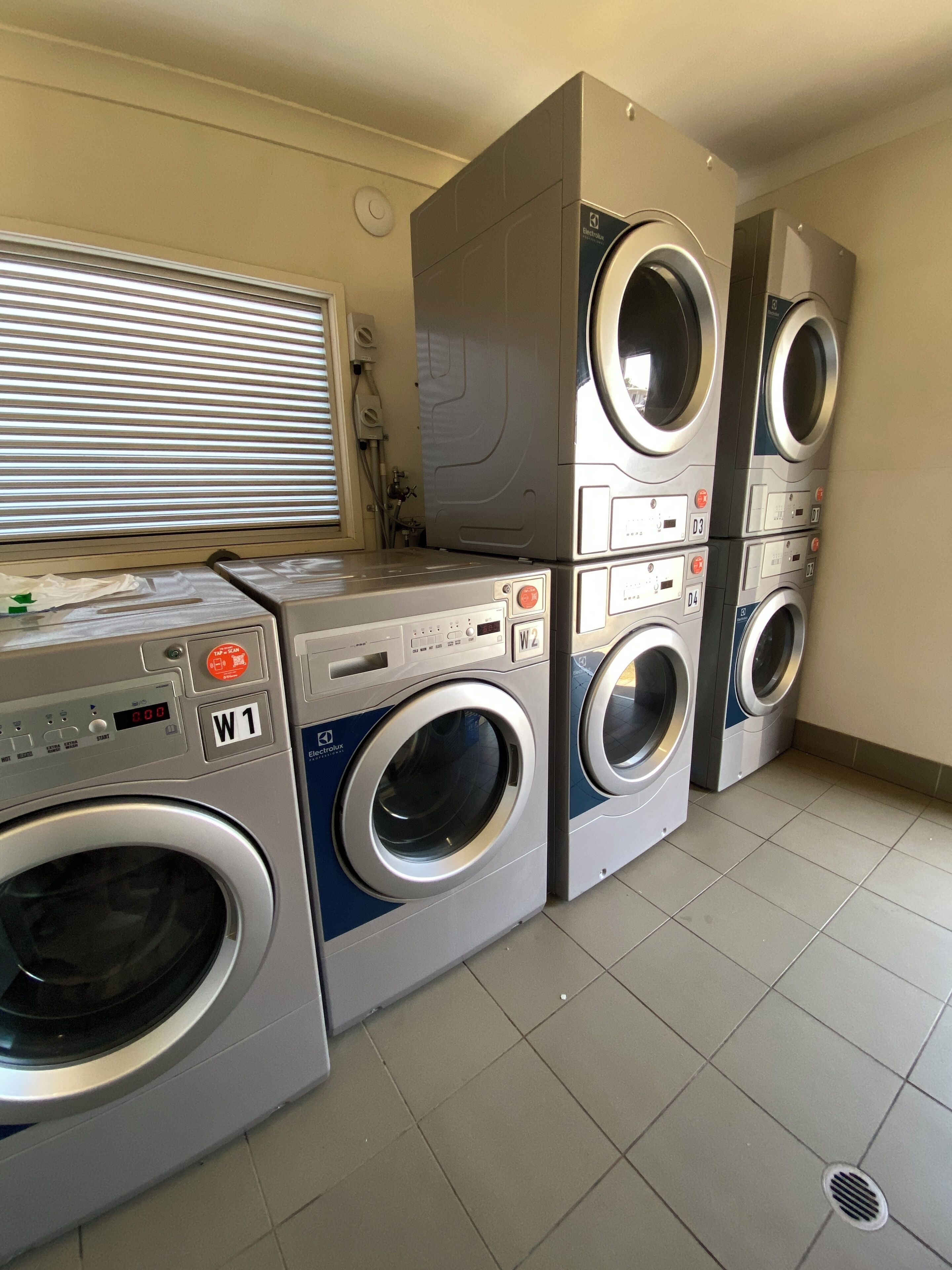 A laundry room with four stacked washing machines, a control panel, and a window with blinds.