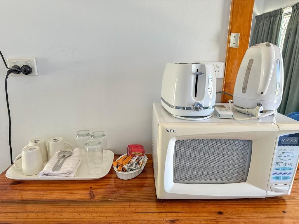 A microwave oven with a digital display and a kettle on top, a tray with cups, glasses, and a spoon, and a power outlet with a cable plugged in.