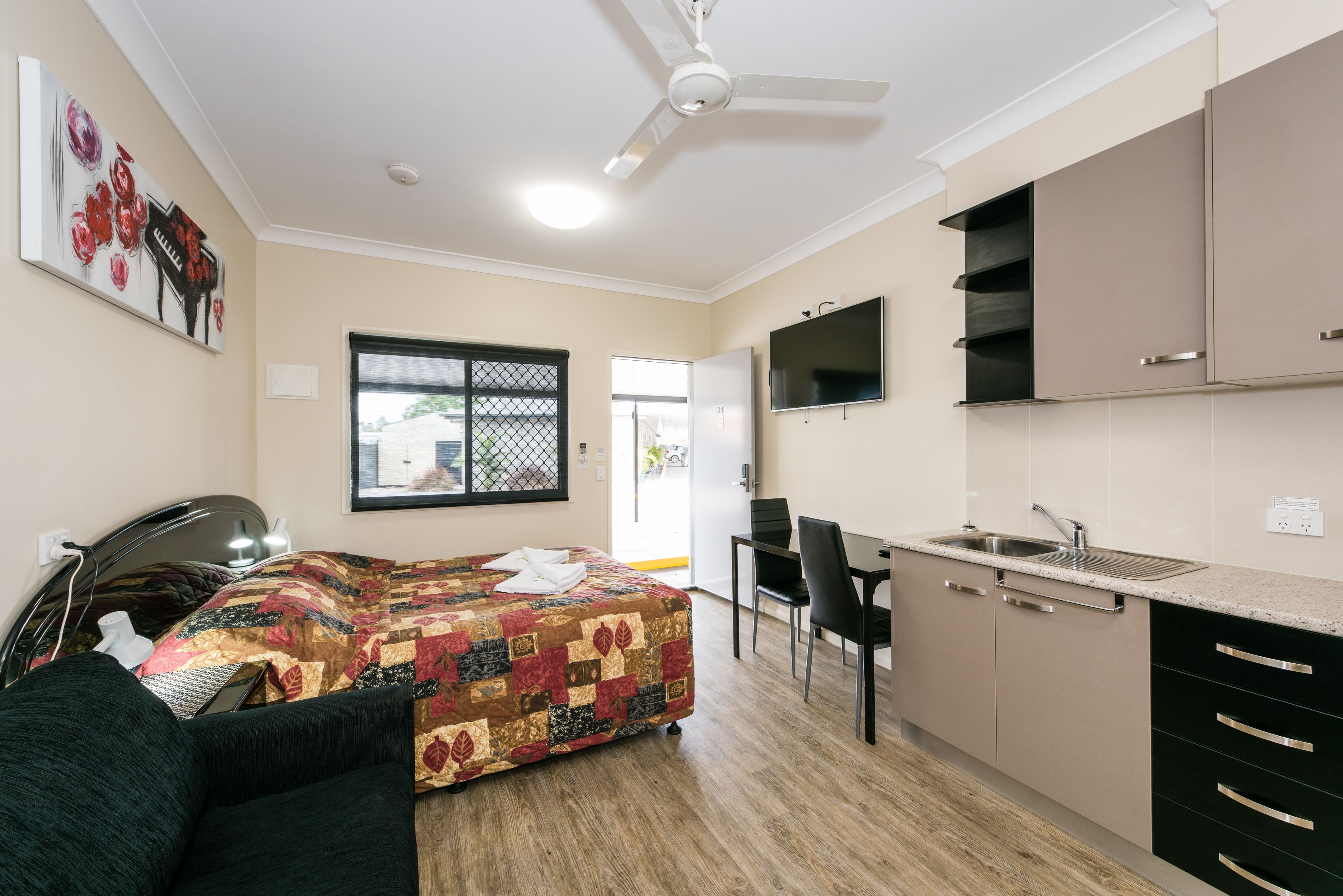 Habitación de hotel compacta con cama, escritorio, kitchenette y ventilador de techo.