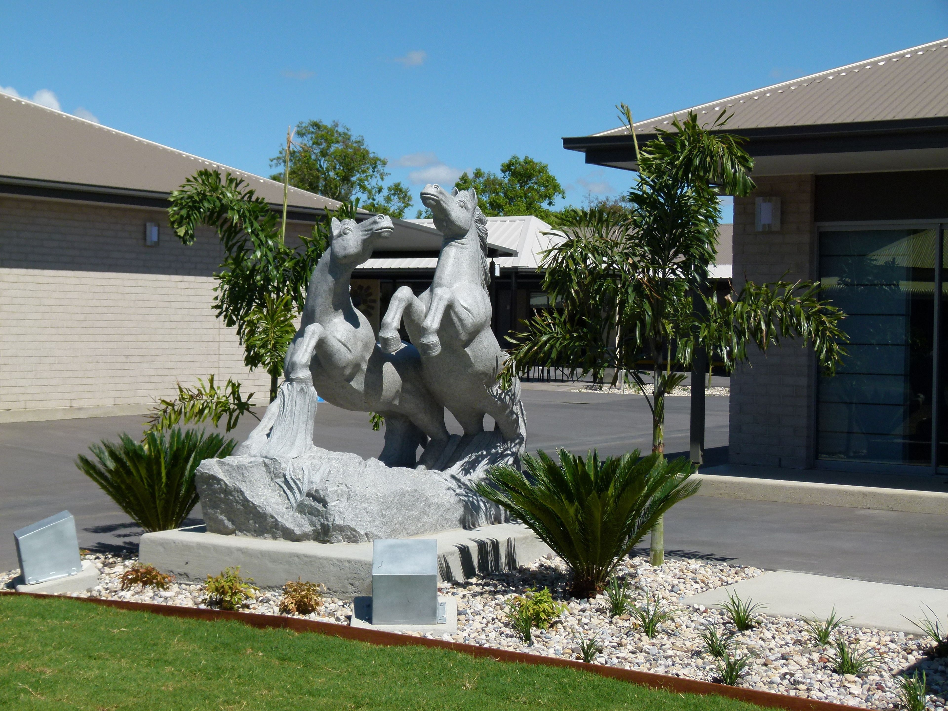 A sculpture of a horse and rider in a landscaped area with palm trees and a building in the background.