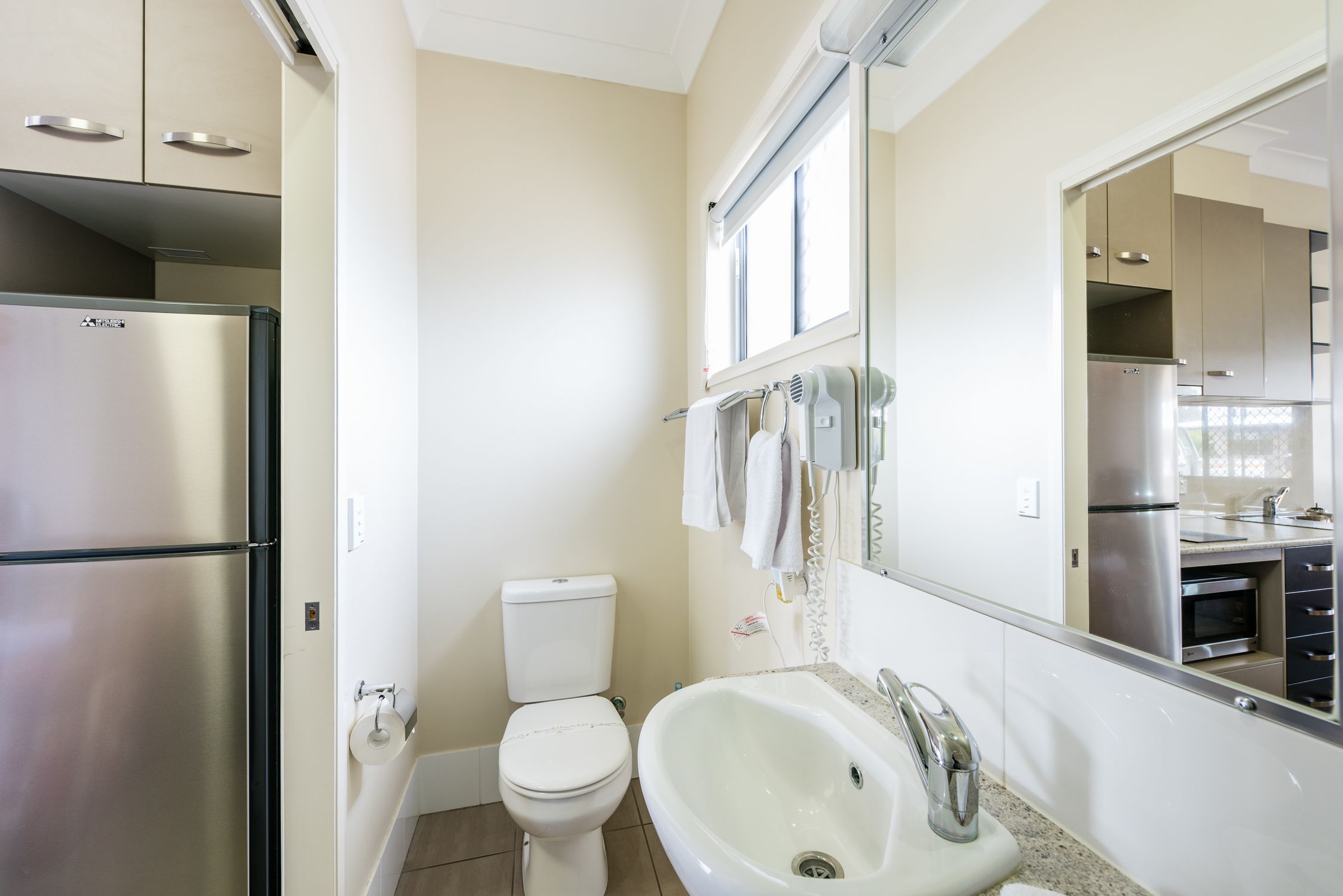 A modern bathroom with a stainless steel refrigerator, a toilet, a sink, and a large mirror.